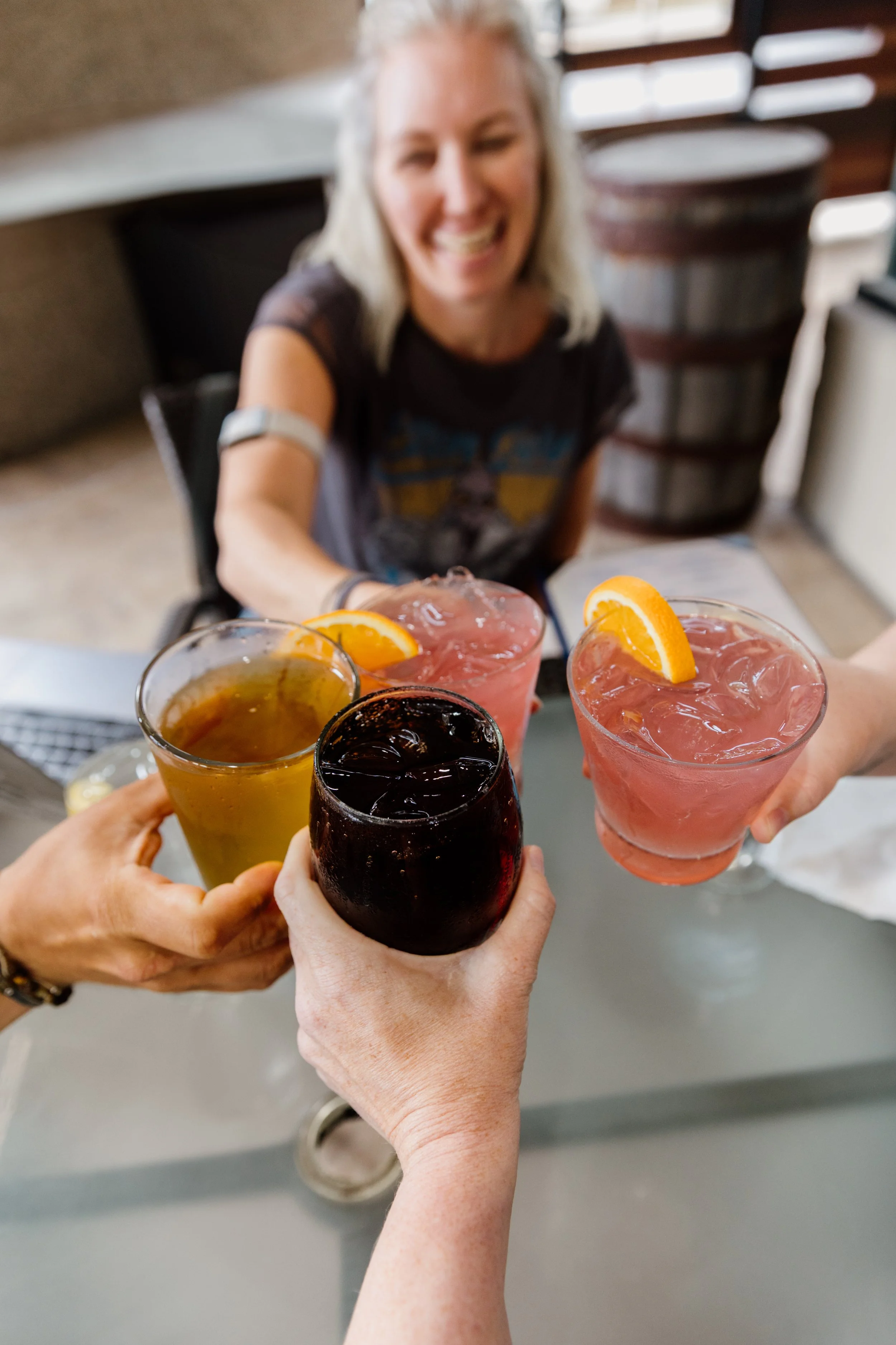 People toasting with drinks, including one with a yellow-colored beverage and others with pink drinks garnished with orange slices, indoor setting.