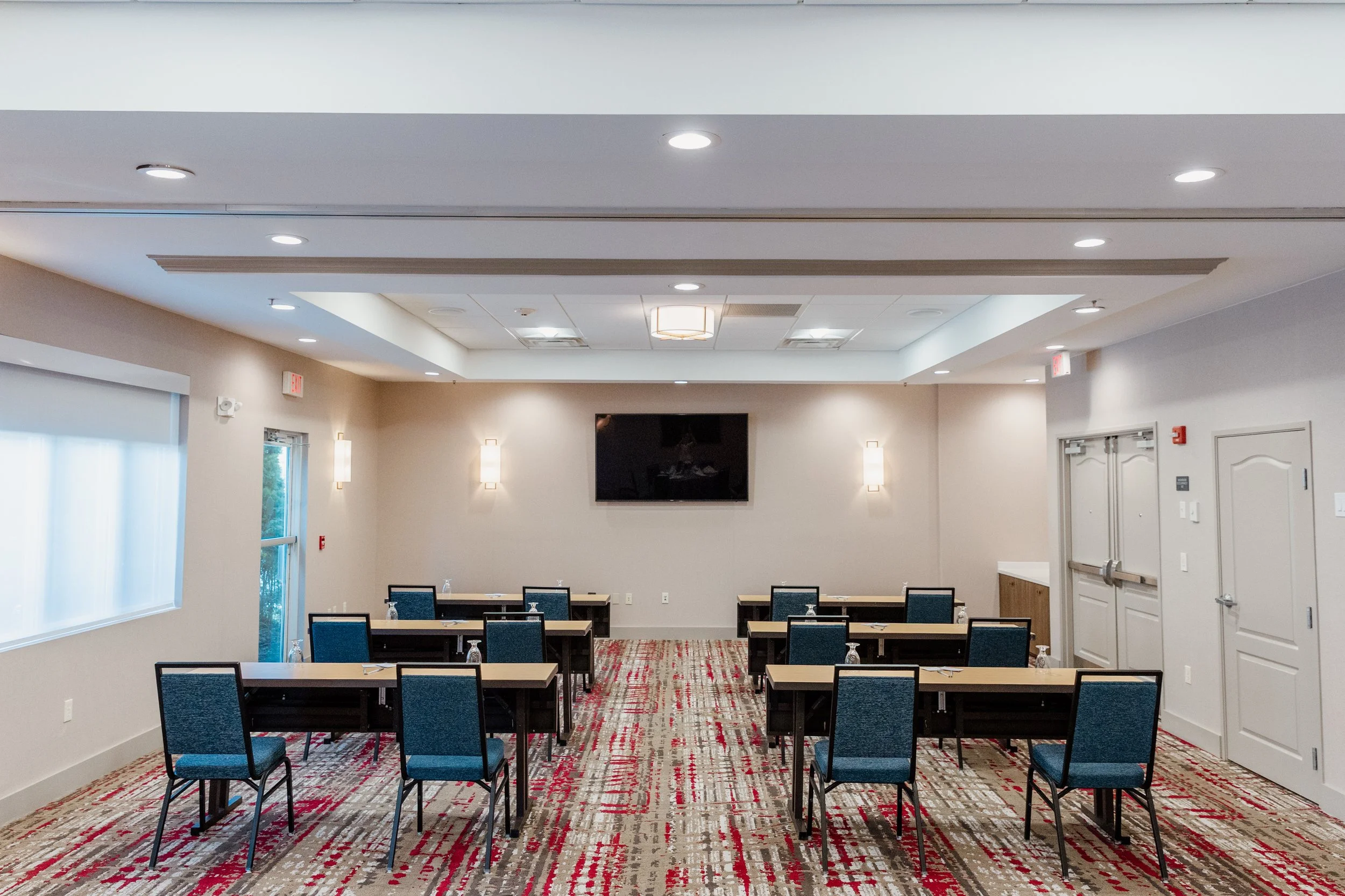 Empty conference room with 8 tables and chairs arranged in two rows. Wall-mounted TV at the front, with beige walls, modern lighting, and a patterned carpet.