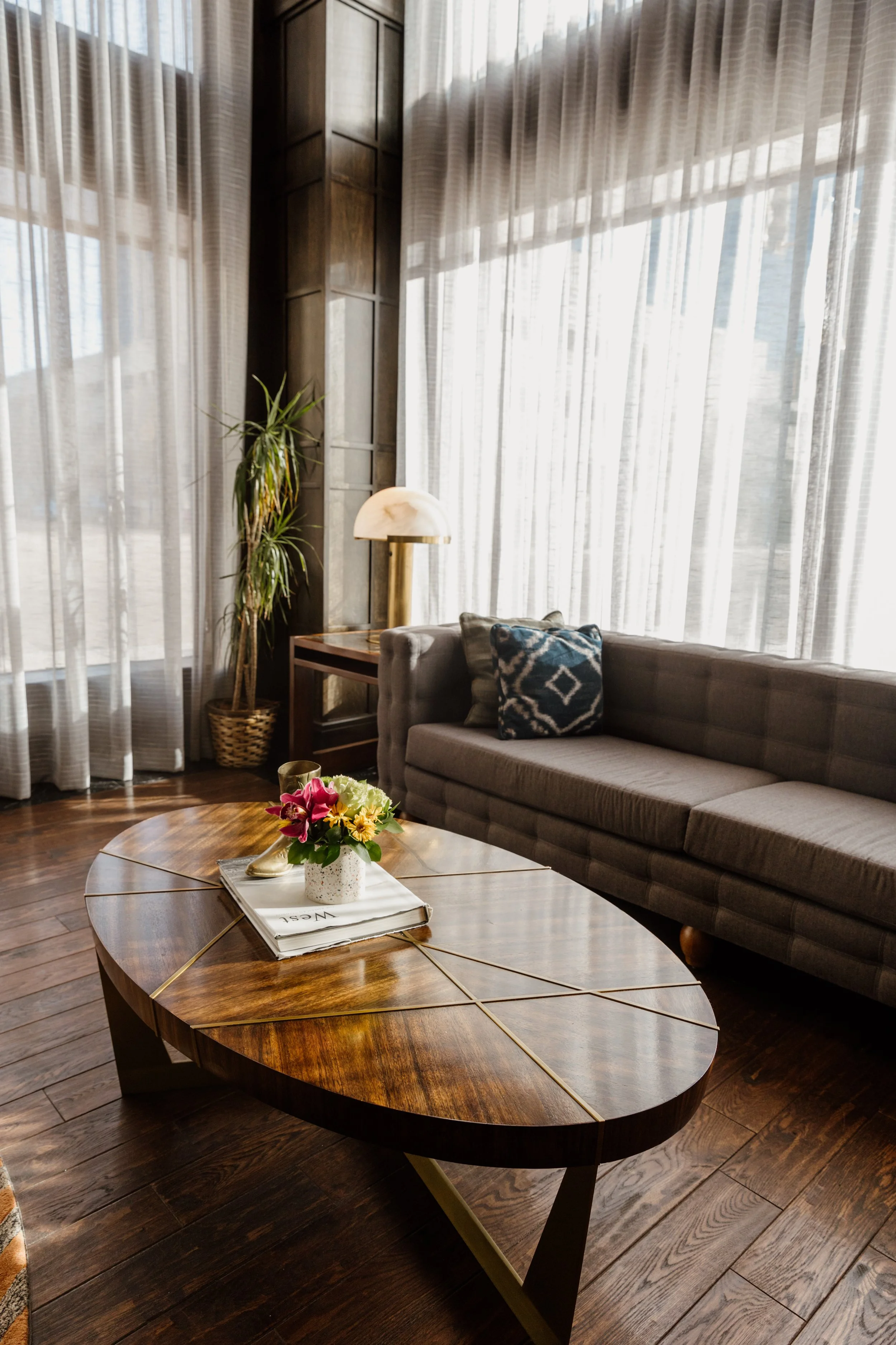 Living room with a dark gray sofa, blue patterned pillow, wooden coffee table with a flower arrangement, tall plant, and window with sheer curtains.