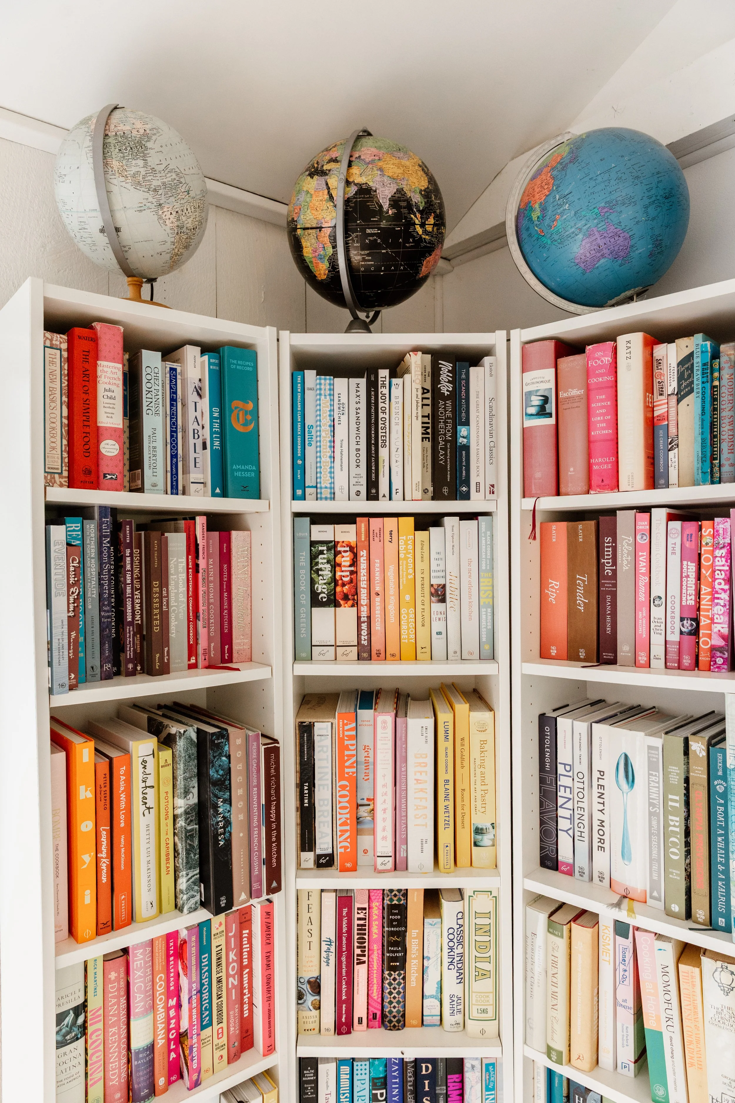 Three globes on top of a white bookshelf filled with colorful books.