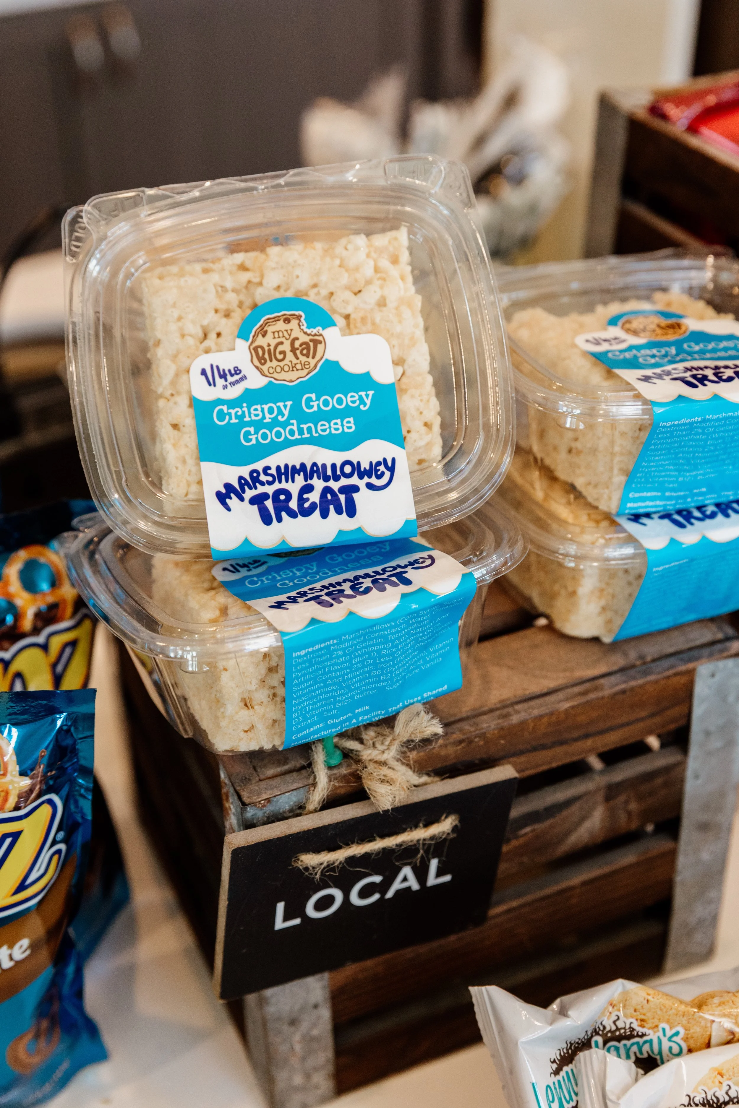 Packages of crispy gooey marshmallow treats labeled as 'Marshmallowy Treat' from 'My Big Fit Cookie,' stacked on a wooden display with a small black sign that says 'Local' underneath.