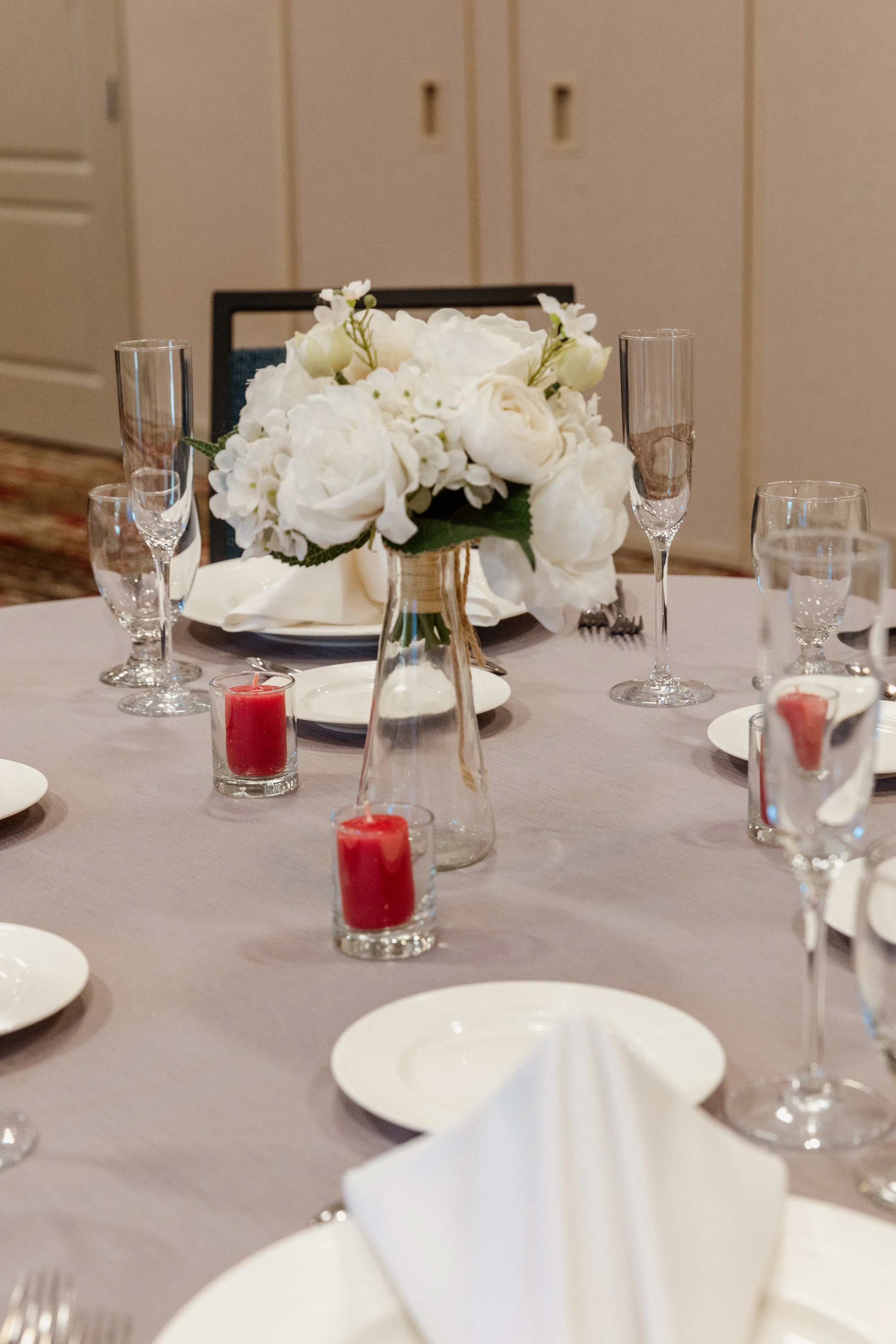 A table set for a formal event with a centerpiece of white flowers in a clear glass vase, red candles in small glass holders, and white plates, napkins, and glassware.