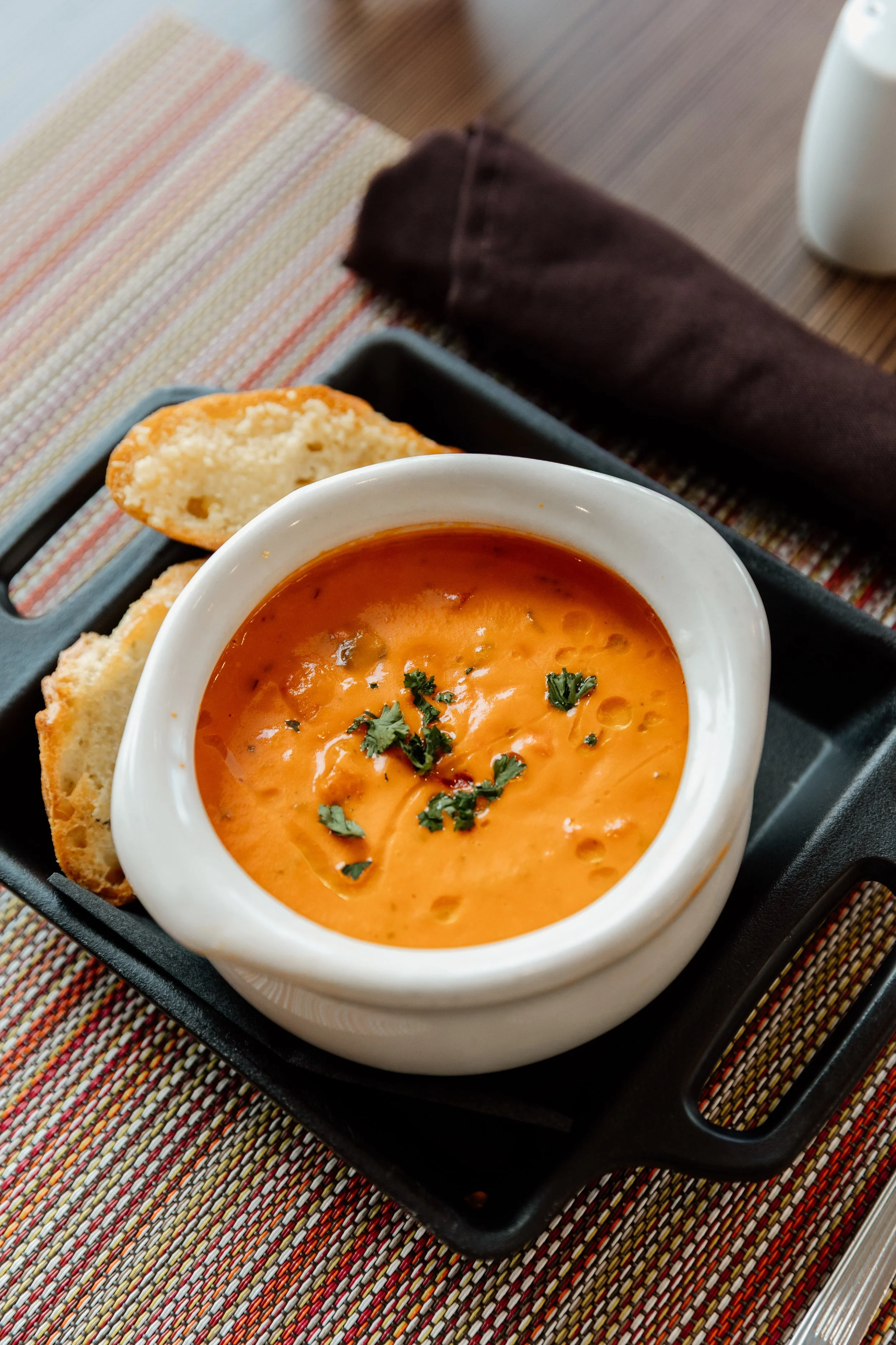 Bowl of creamy tomato soup garnished with herbs, served with slices of bread on a black tray, placed on a colorful striped tablecloth.