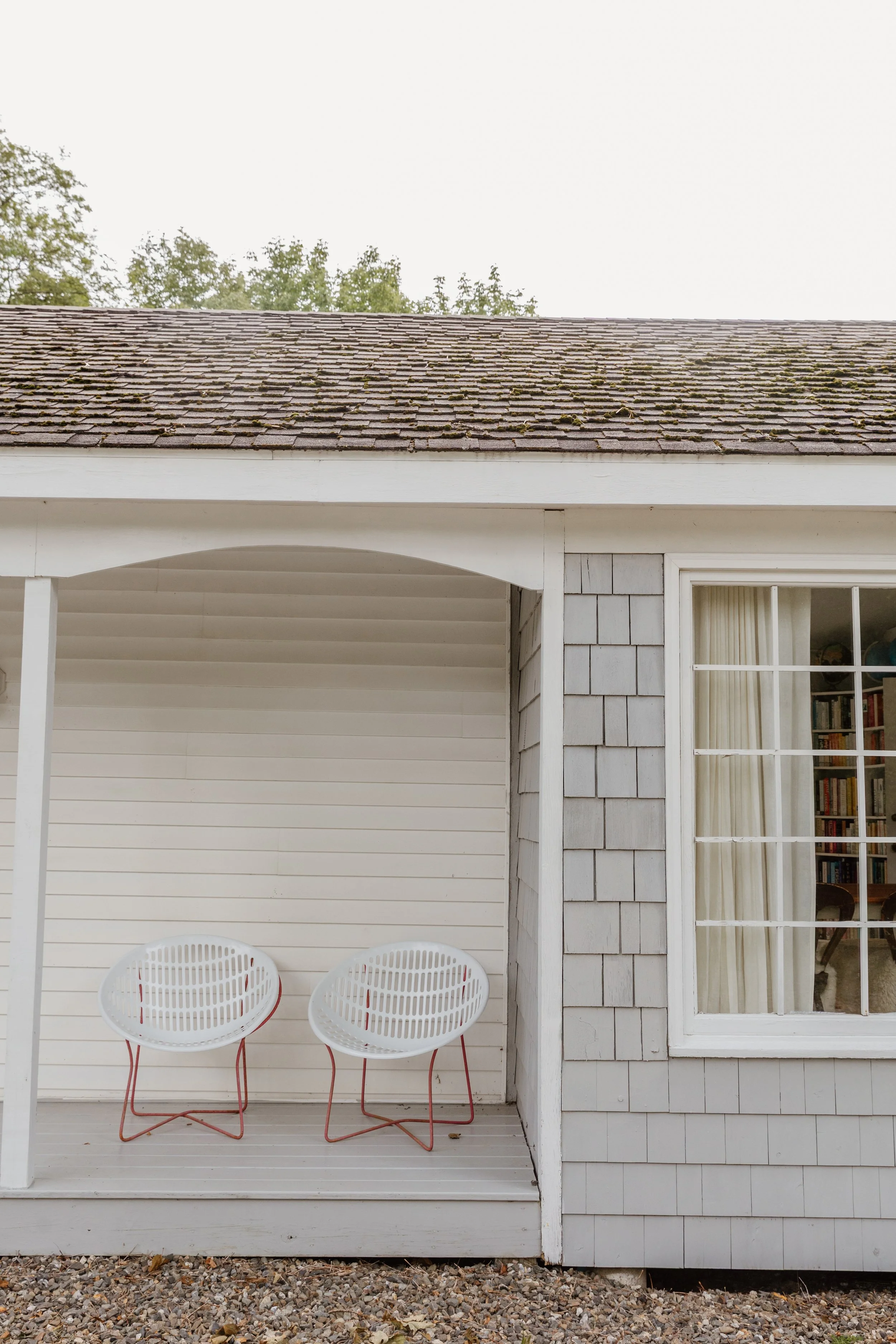 Two white chairs with red legs on a porch in front of a window and house siding.