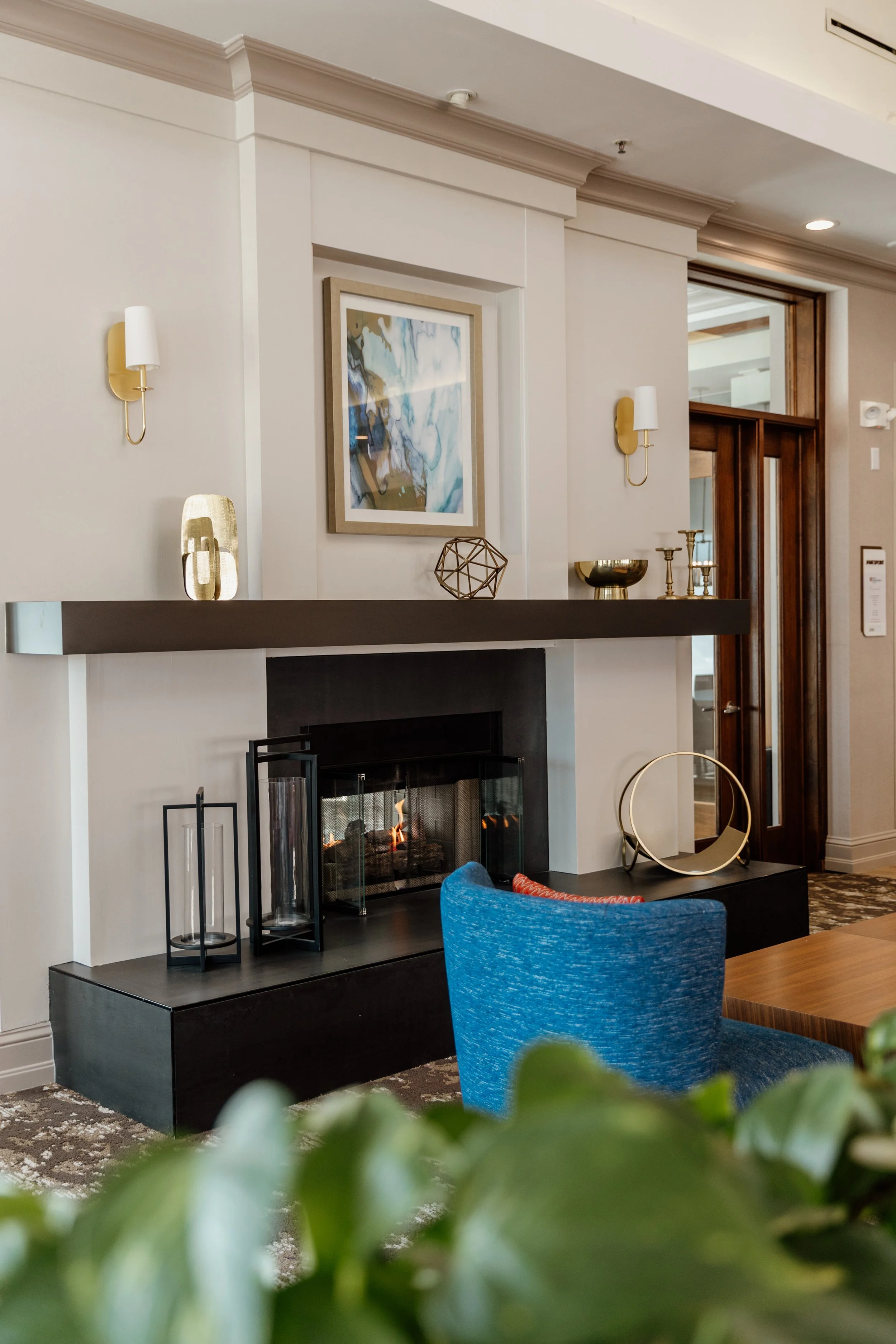 Modern fireplace in a hotel lobby with decorative objects on the mantel, framed artwork above, and a blue chair in the foreground.