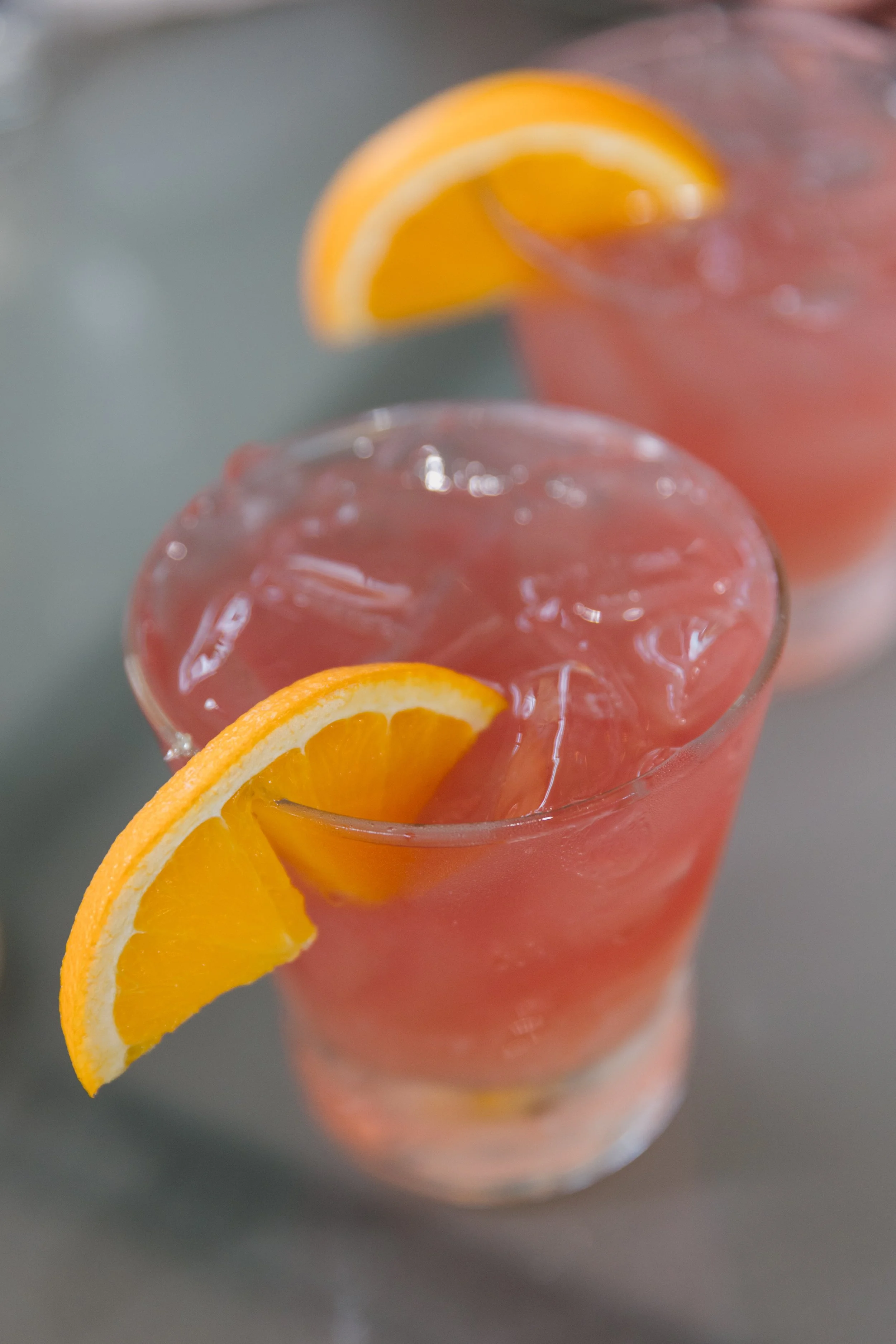 Two glasses of pink lemonade with ice and orange wedges on top, placed on a gray surface.
