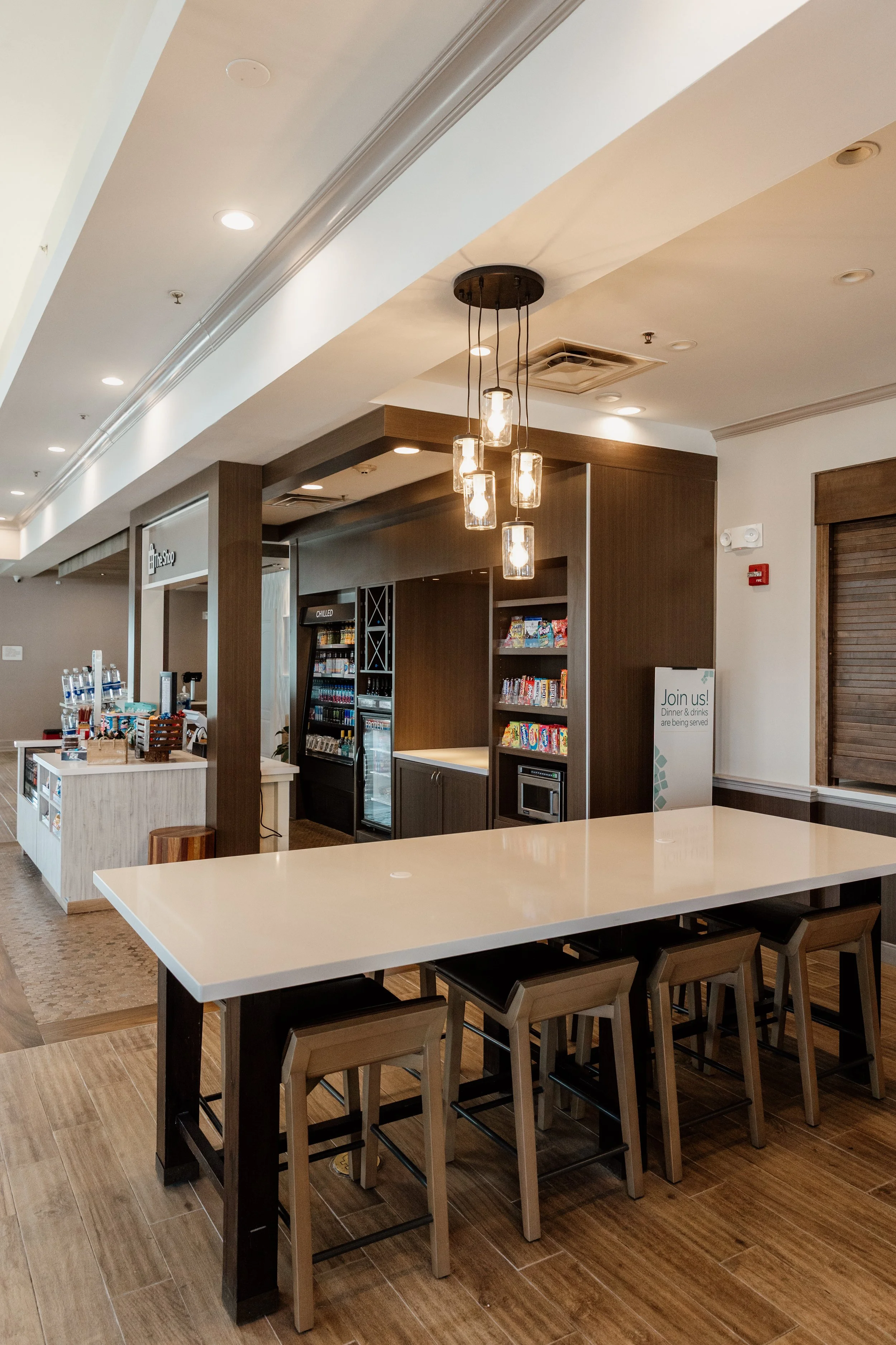 Interior of a cafe or dining area with a beige table and four bar stools, dark wood shelves, a vending machine, and a sign that says "Join us! Dinner & drinks are being served."