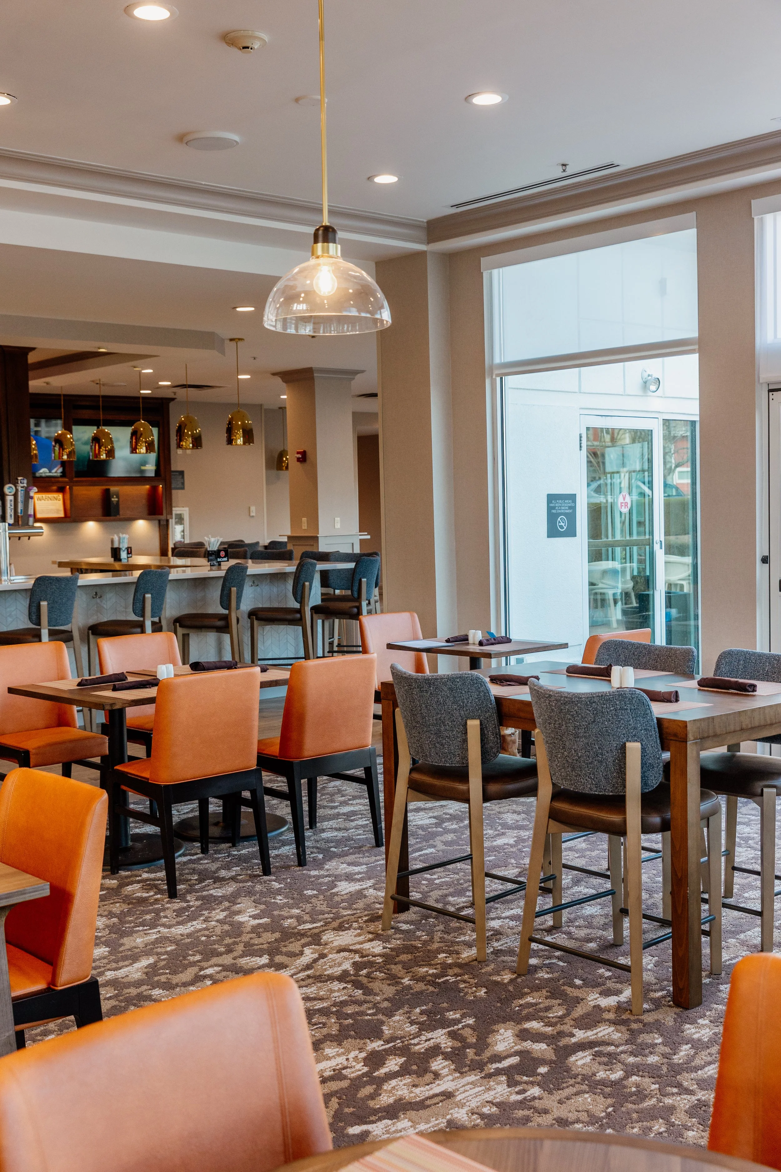 Empty restaurant dining area with tables and chairs, bar counter in the background, large windows, and ceiling lights.