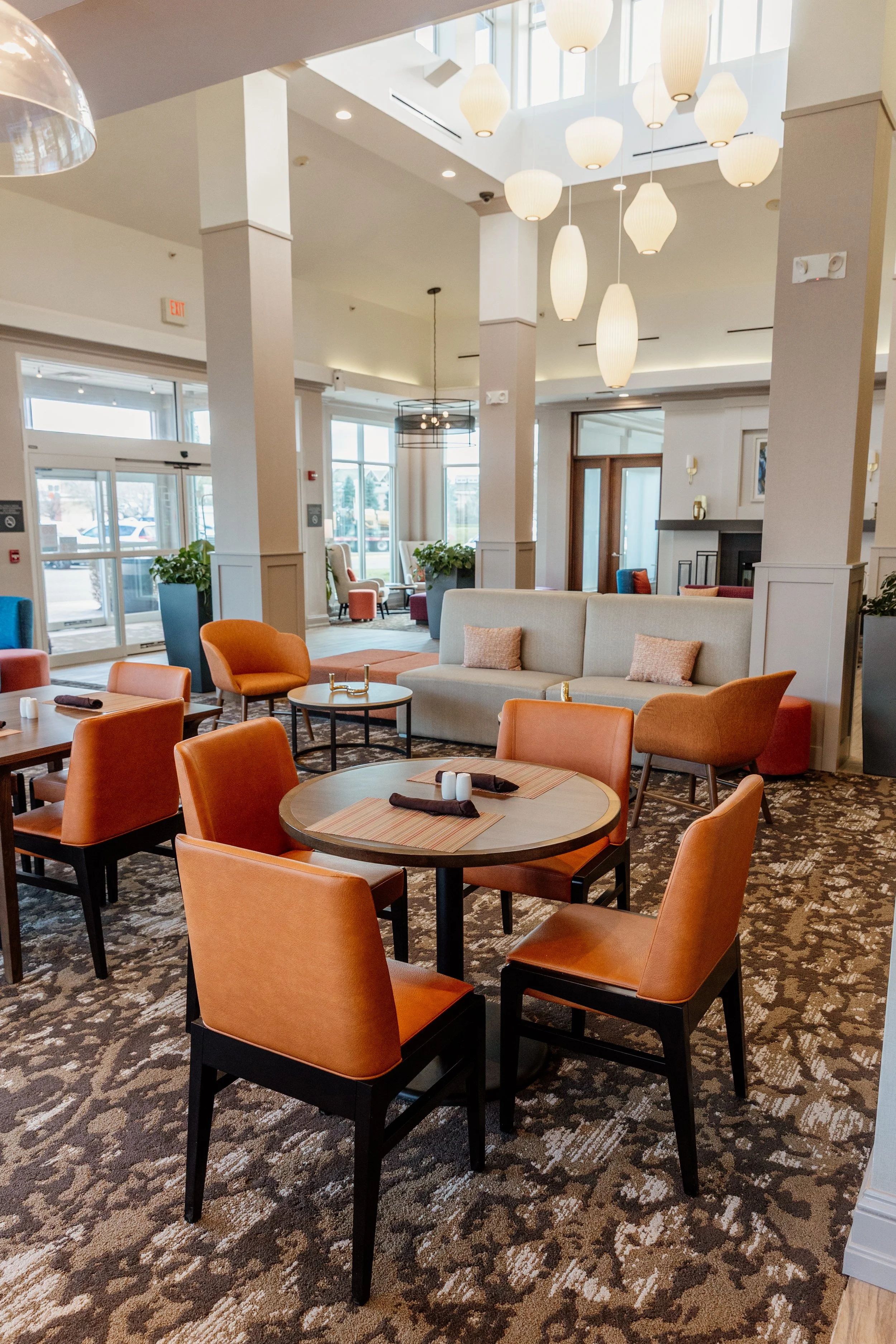 Elegant restaurant or lounge with warm lighting, orange chairs, round tables with place settings, white sofas with pillows, and large windows letting in natural light.