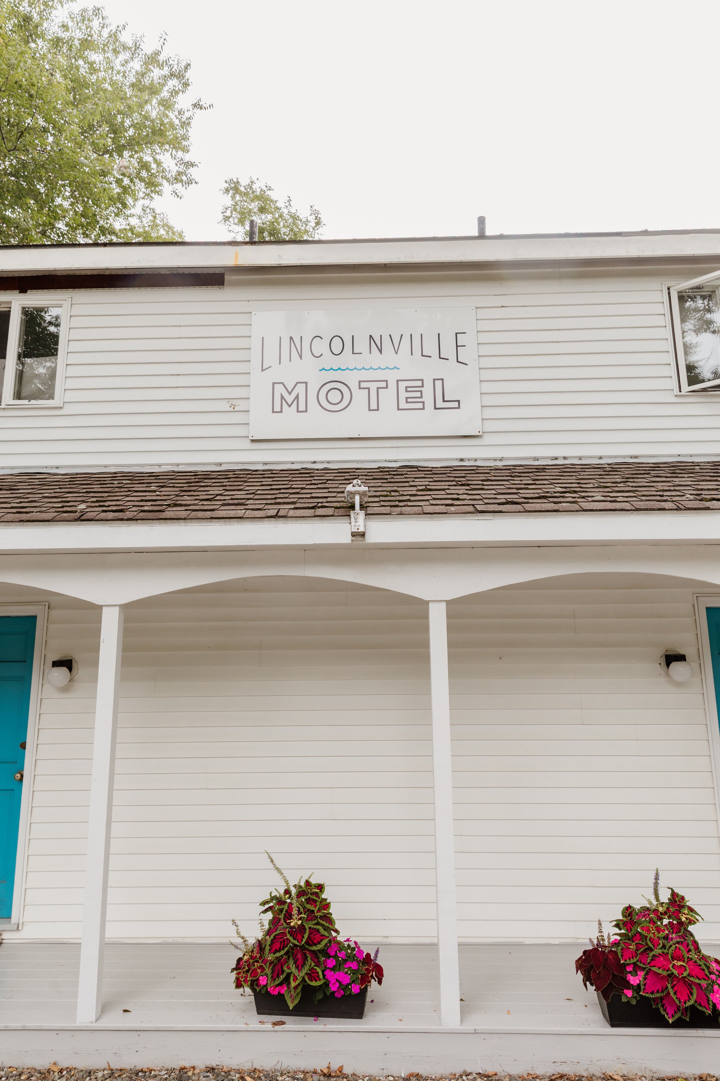 The charming exterior of the Lincolnville Motel in Lincolnville, Maine, with its white facade, signature turquoise doors, and cheerful flower pots out front.