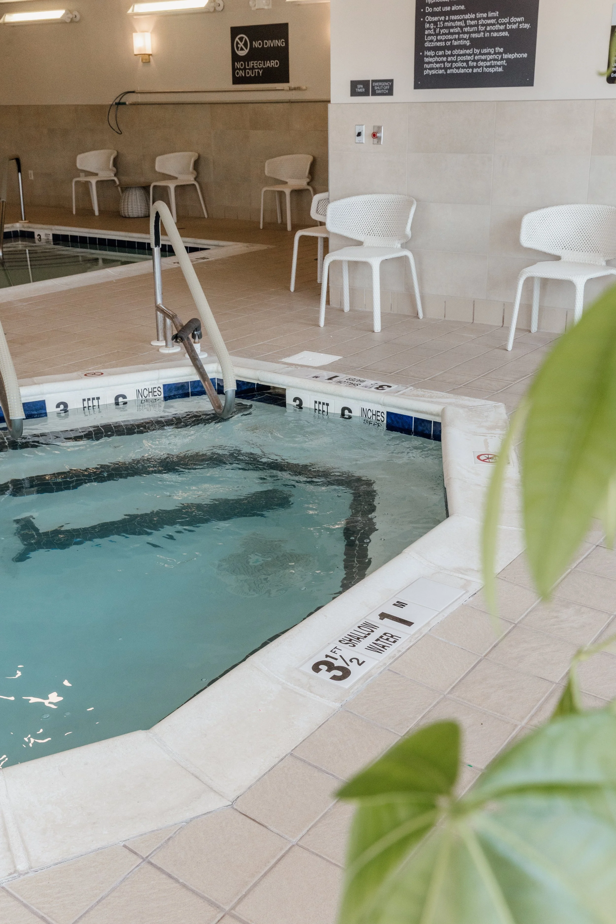 Indoor swimming pool area with white chairs near the edge, safety and depth signs, and a no diving sign on the wall.