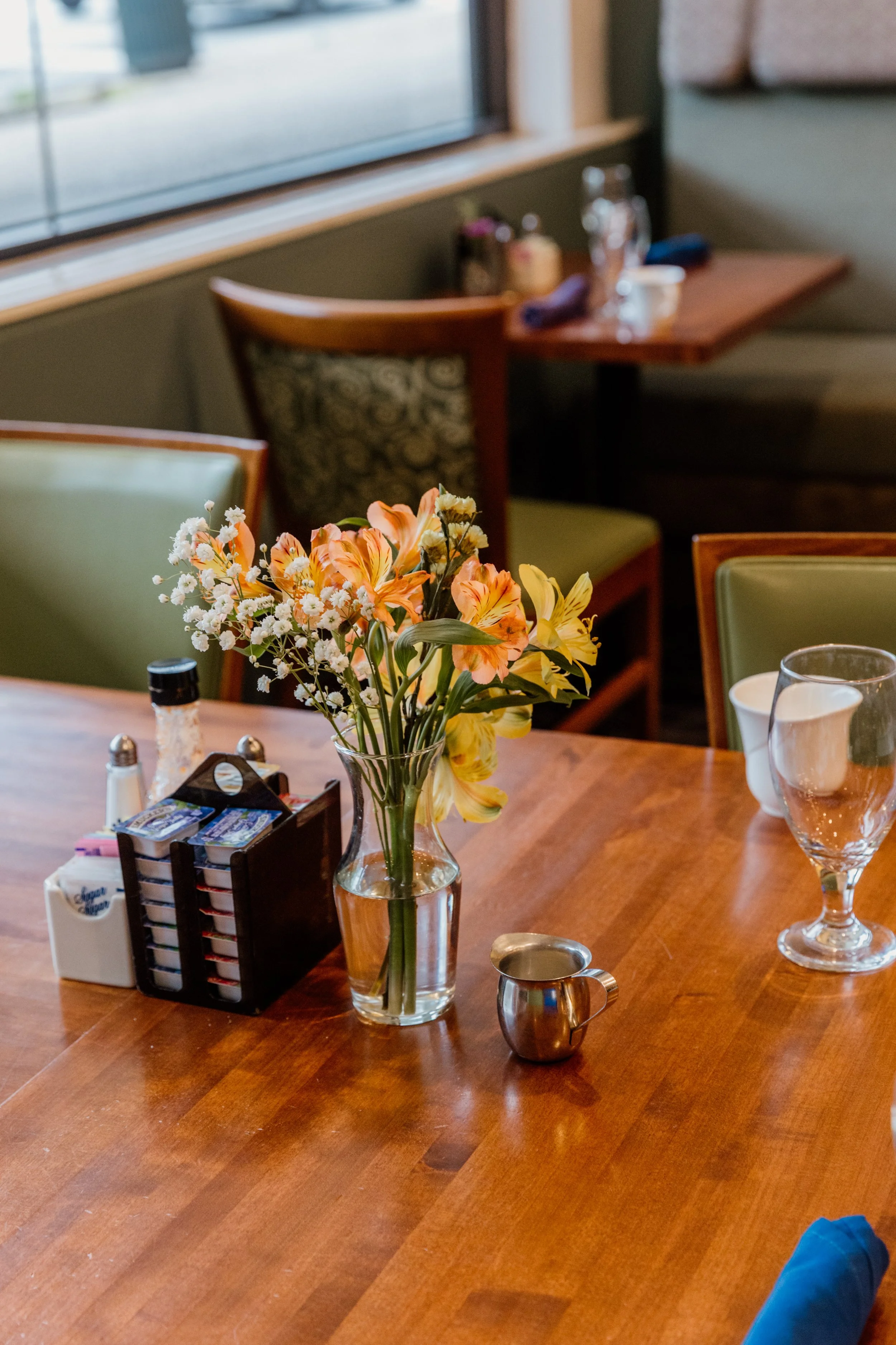A wooden dining table with a vase of yellow and pink flowers, transfer sugar, salt and pepper shakers, a napkin holder, a glass water pitcher, and a empty water glass in a restaurant setting.