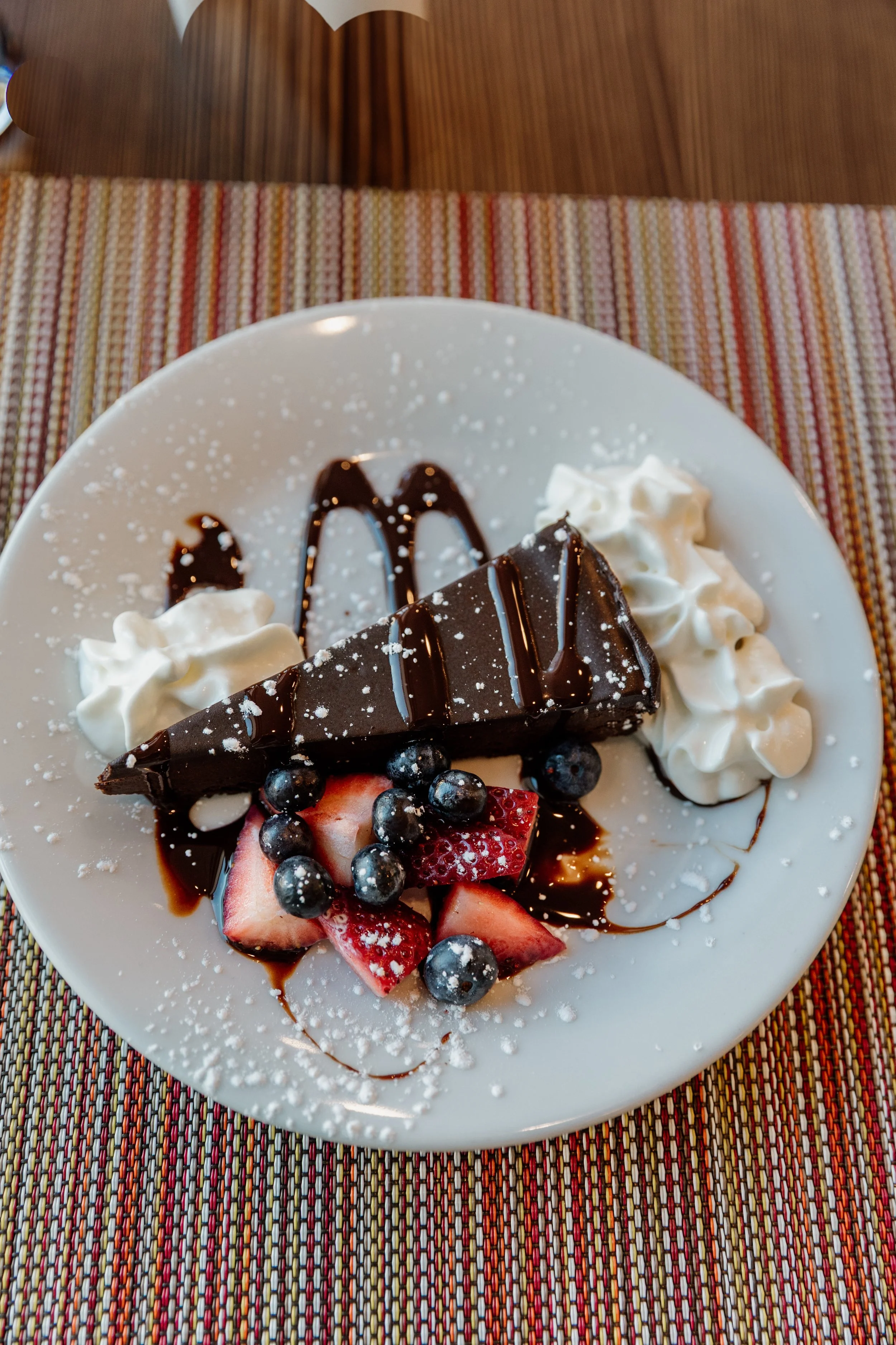 Slice of chocolate cheesecake with whipped cream, blueberries, strawberries, chocolate drizzle, and powdered sugar on a white plate with a colorful tablecloth.