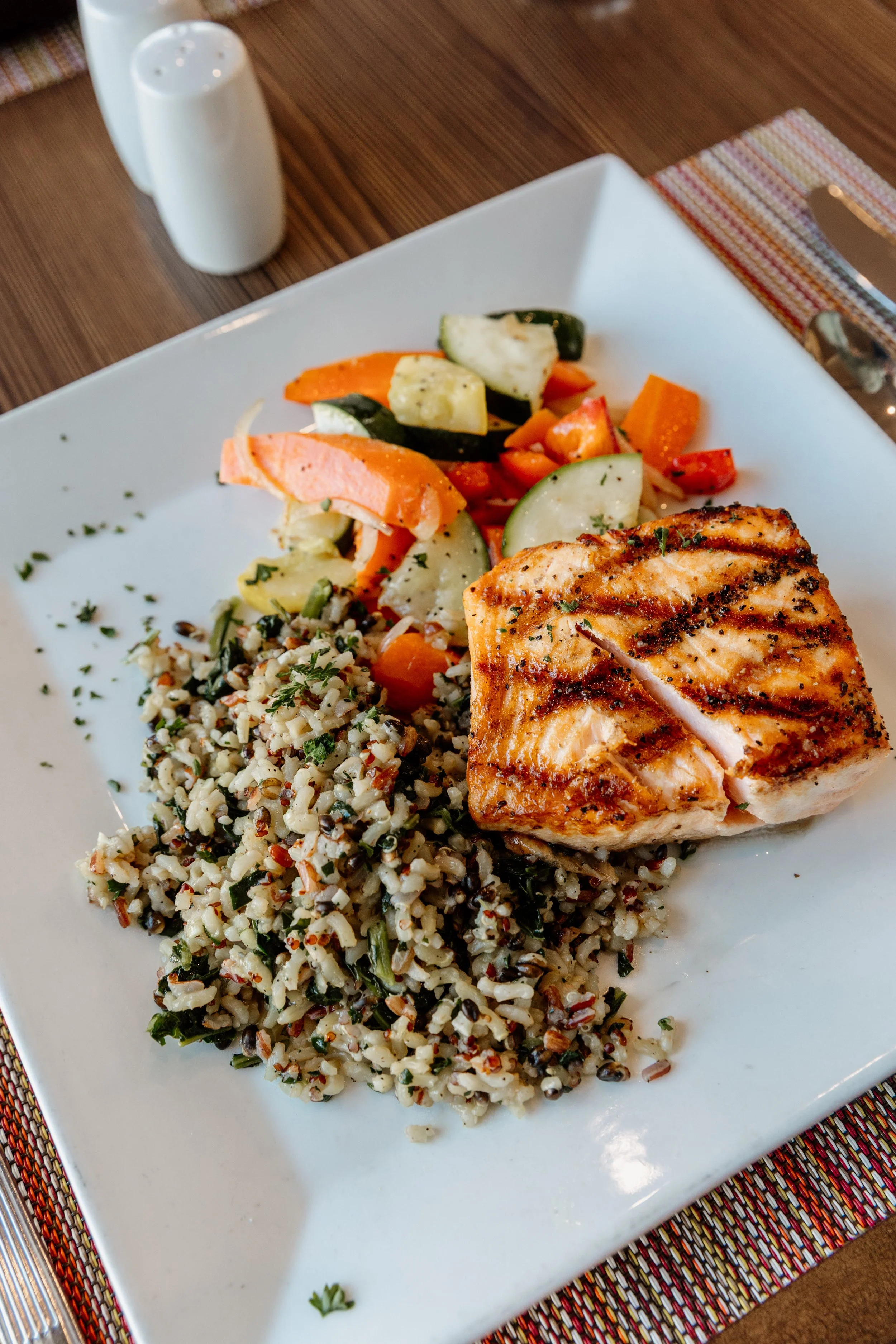 Grilled salmon with dark grill marks, served with a side of rice and chopped vegetables including carrots, zucchini, and peppers.