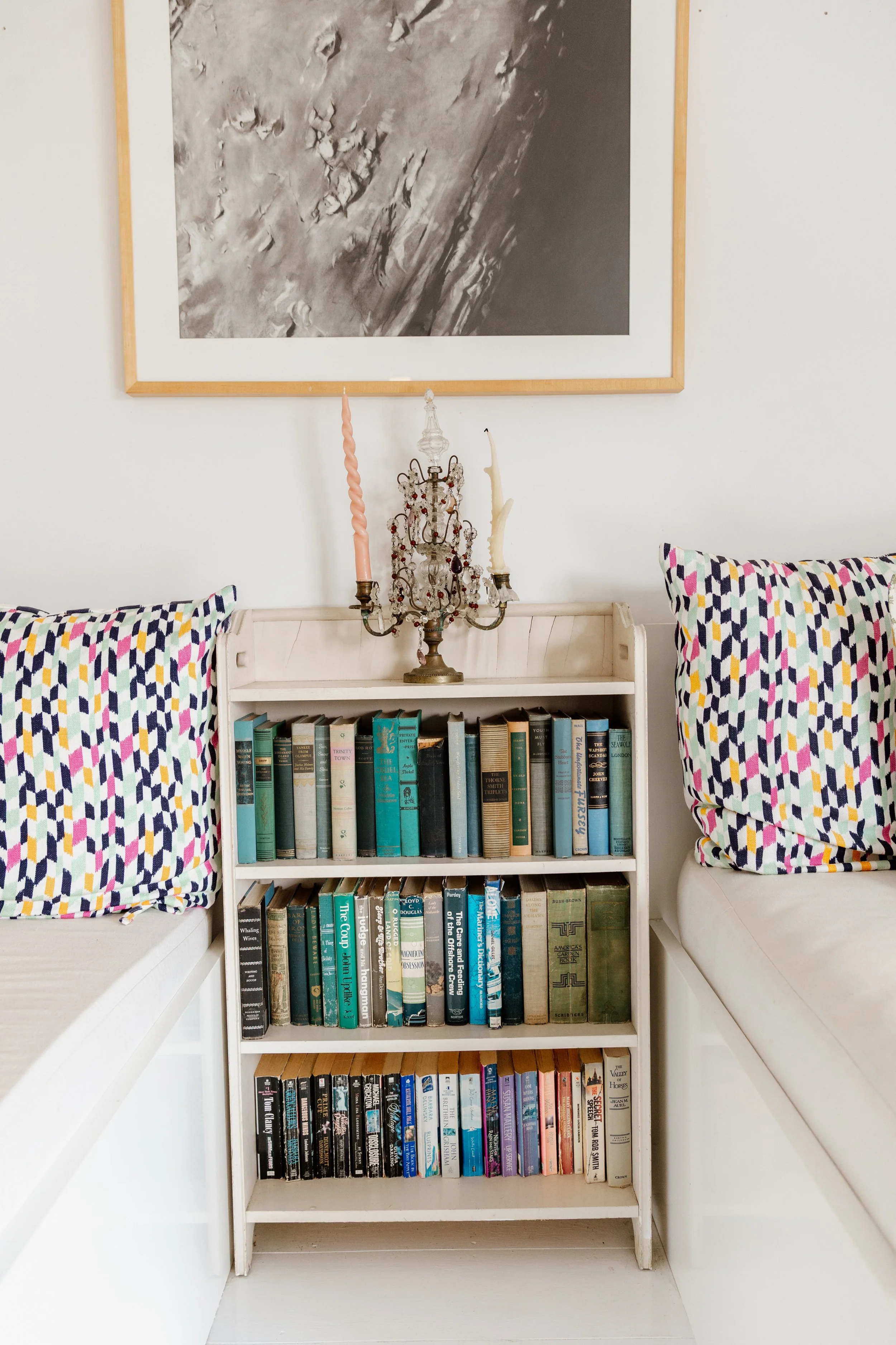 A white bookshelf filled with books, situated between two beds with colorful patterned pillows. A vintage chandelier with candles is placed on top of the bookshelf, and a large framed black-and-white photograph hangs above it.