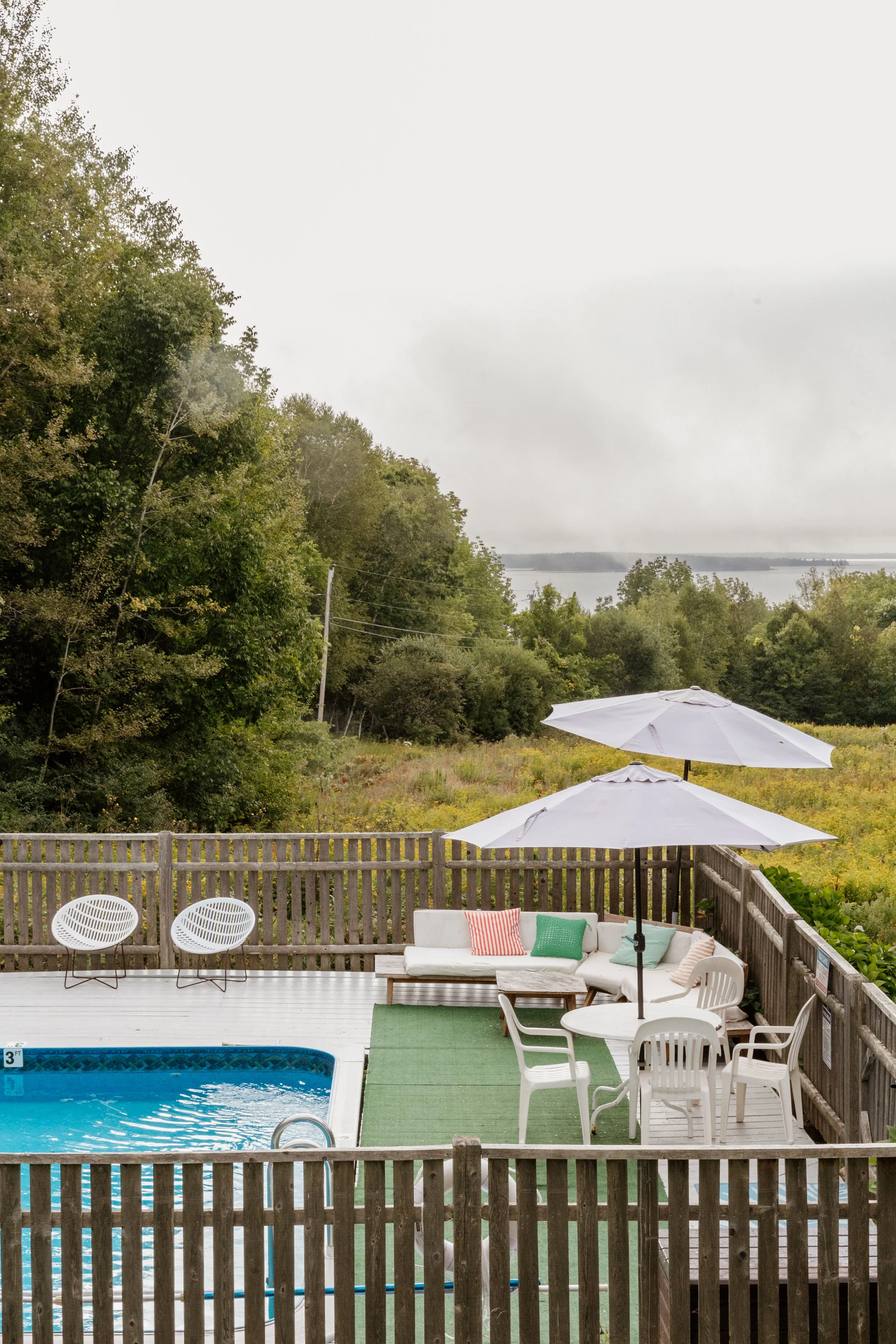 A backyard patio with a swimming pool, white chairs, a table, umbrellas, and a cushioned sitting area with pillows, overlooking a green landscape and water.