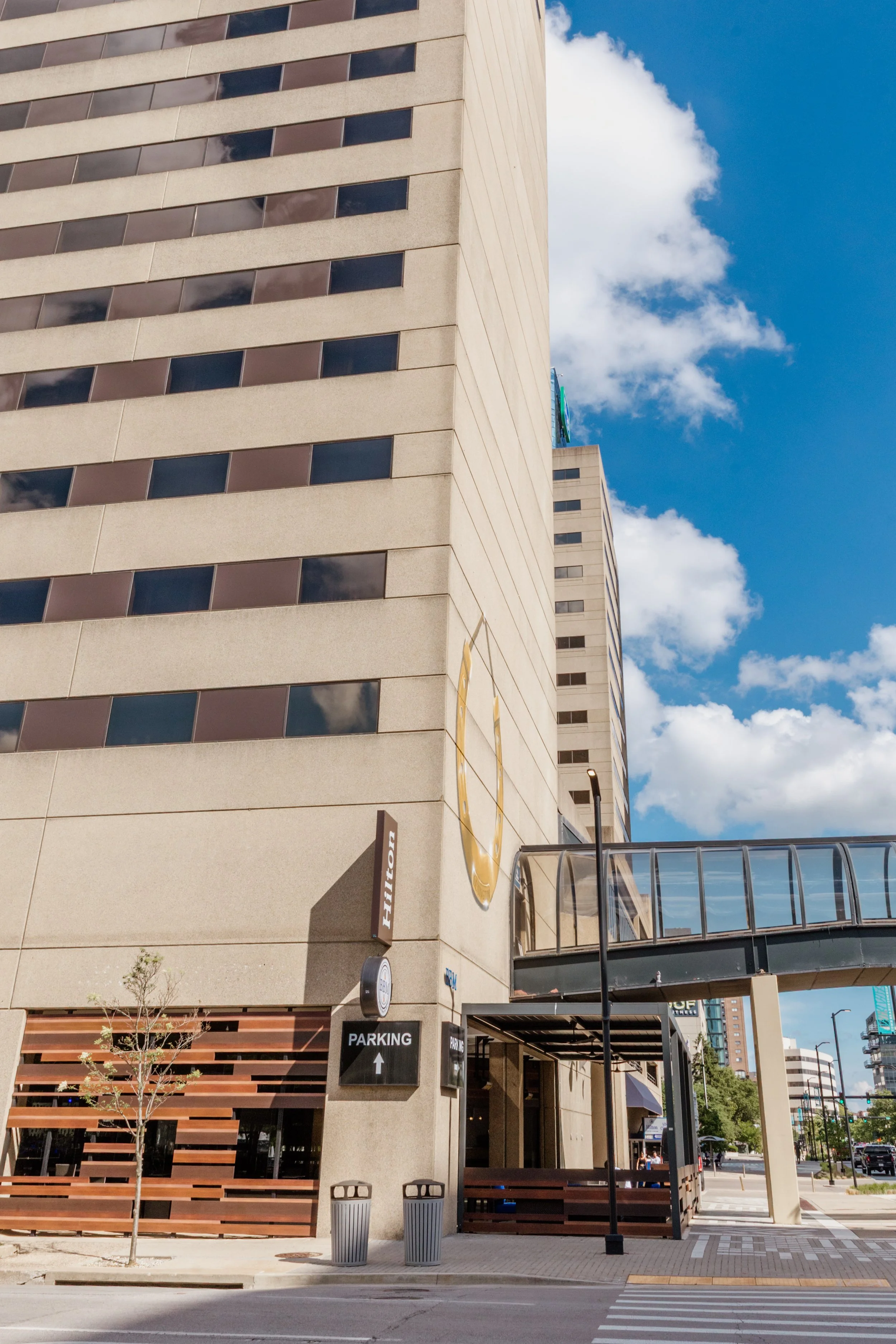 Tall university building with a large emblem and a pedestrian bridge connecting to the sidewalk, under a partly cloudy blue sky.