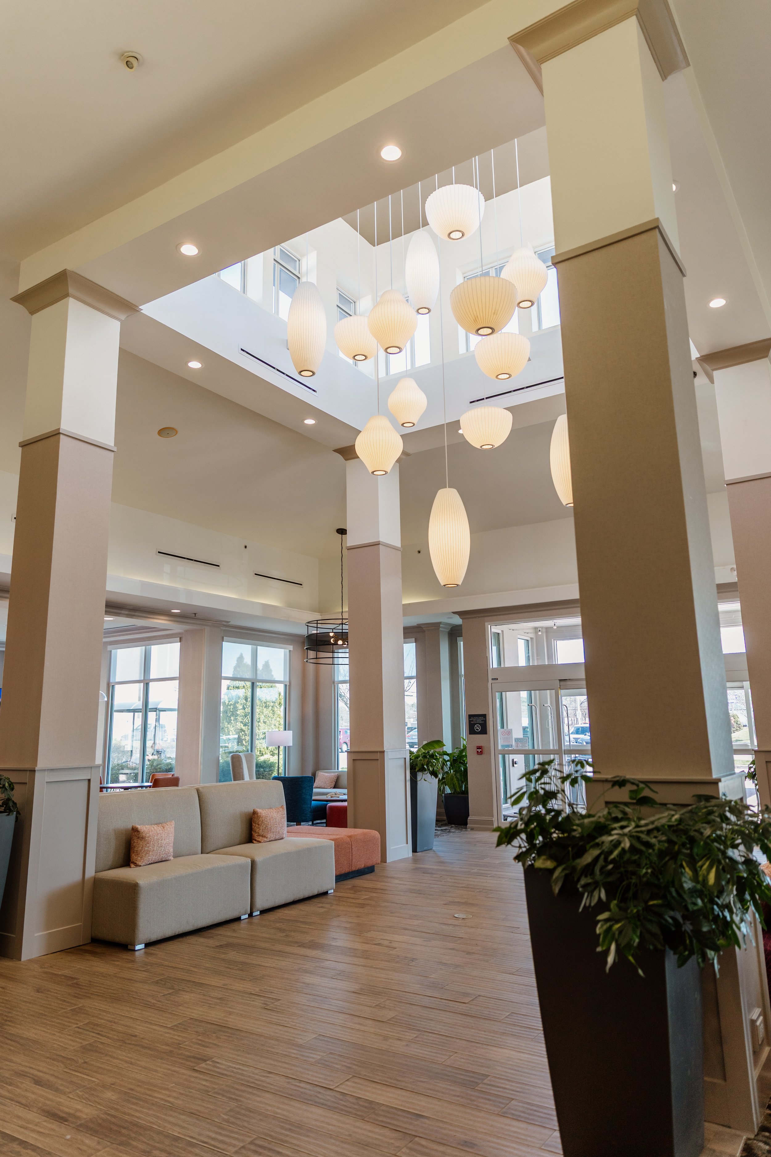 Interior of a modern hotel lobby with high ceilings, paper pendant lighting, large windows, and a beige sofa near potted plants.