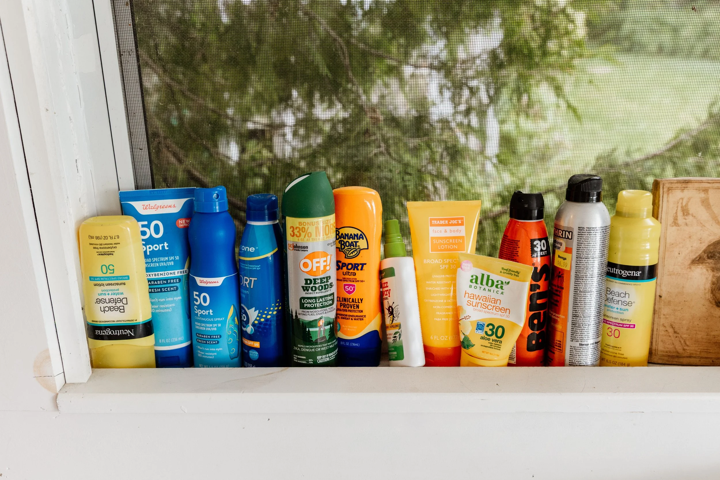 A collection of various sunscreen and bug spray bottles and tubes on a windowsill with a mesh screen and trees outside.