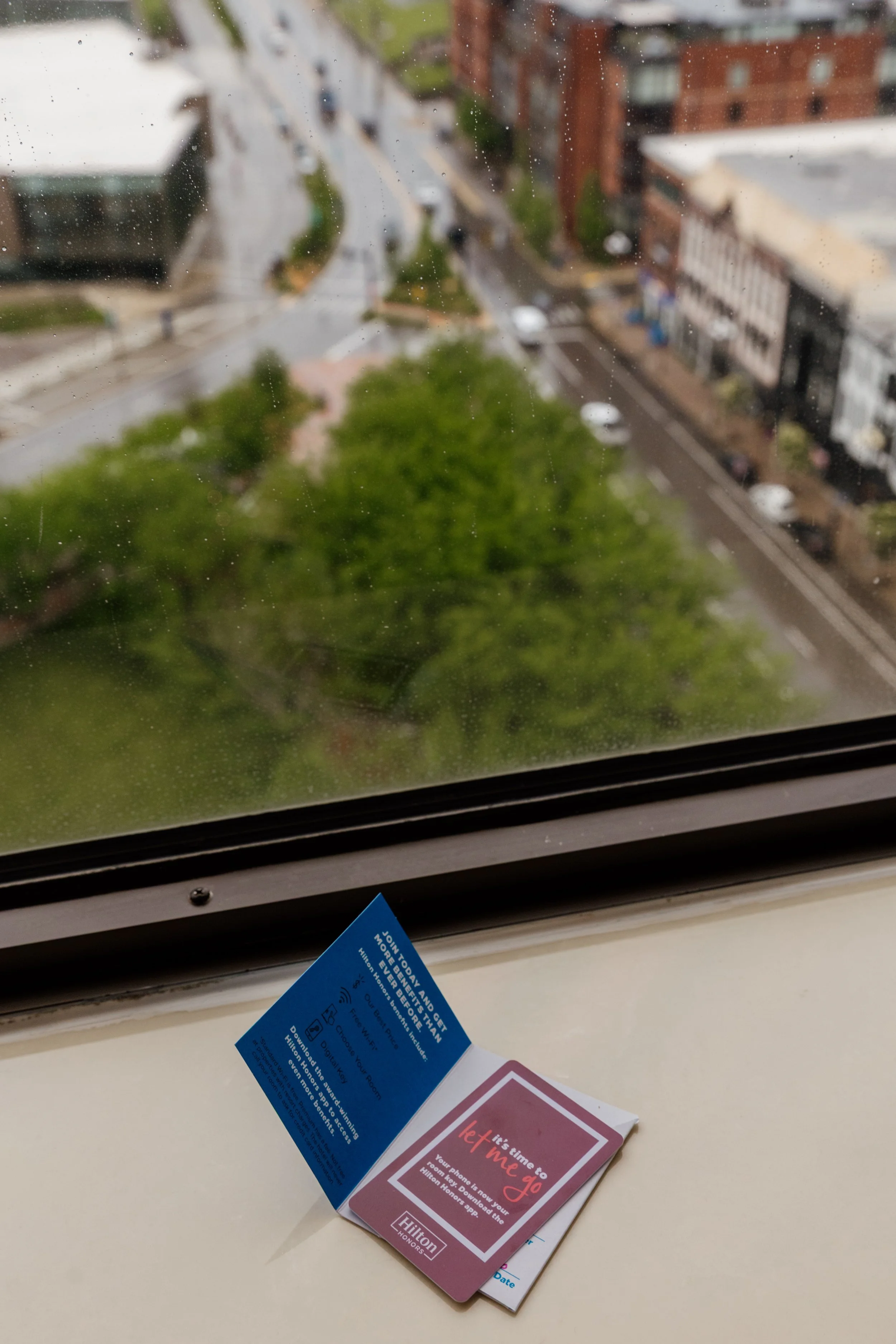 View of a city street through a window with rain droplets, with a blue and pink hotel key card resting on the windowsill in the foreground.