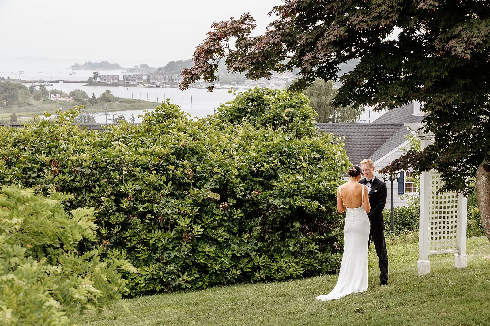 A bride and groom sharing a quiet moment at Haley Mansion in Mystic, Connecticut, the bride in a stunning open back white gown and the groom in a classic black tuxedo, with lush greenery and waterfront views behind them.
