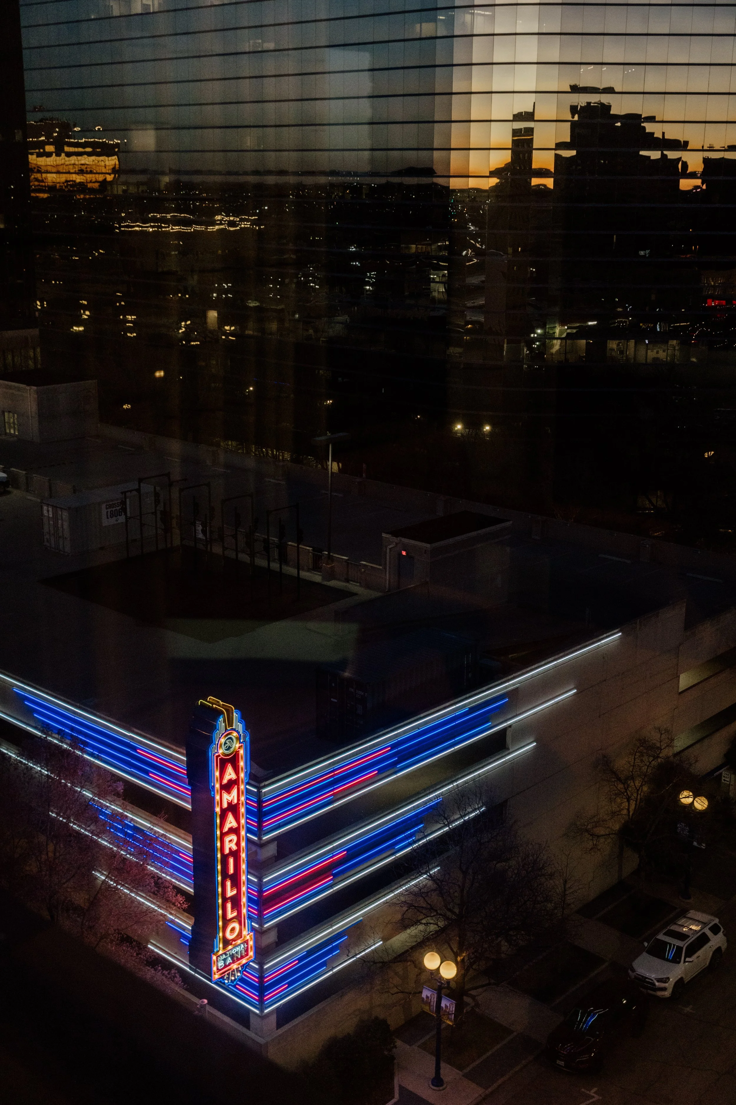 Nighttime cityscape viewed through window with reflection, neon sign reading 'AMARILLO,' and streetlights.