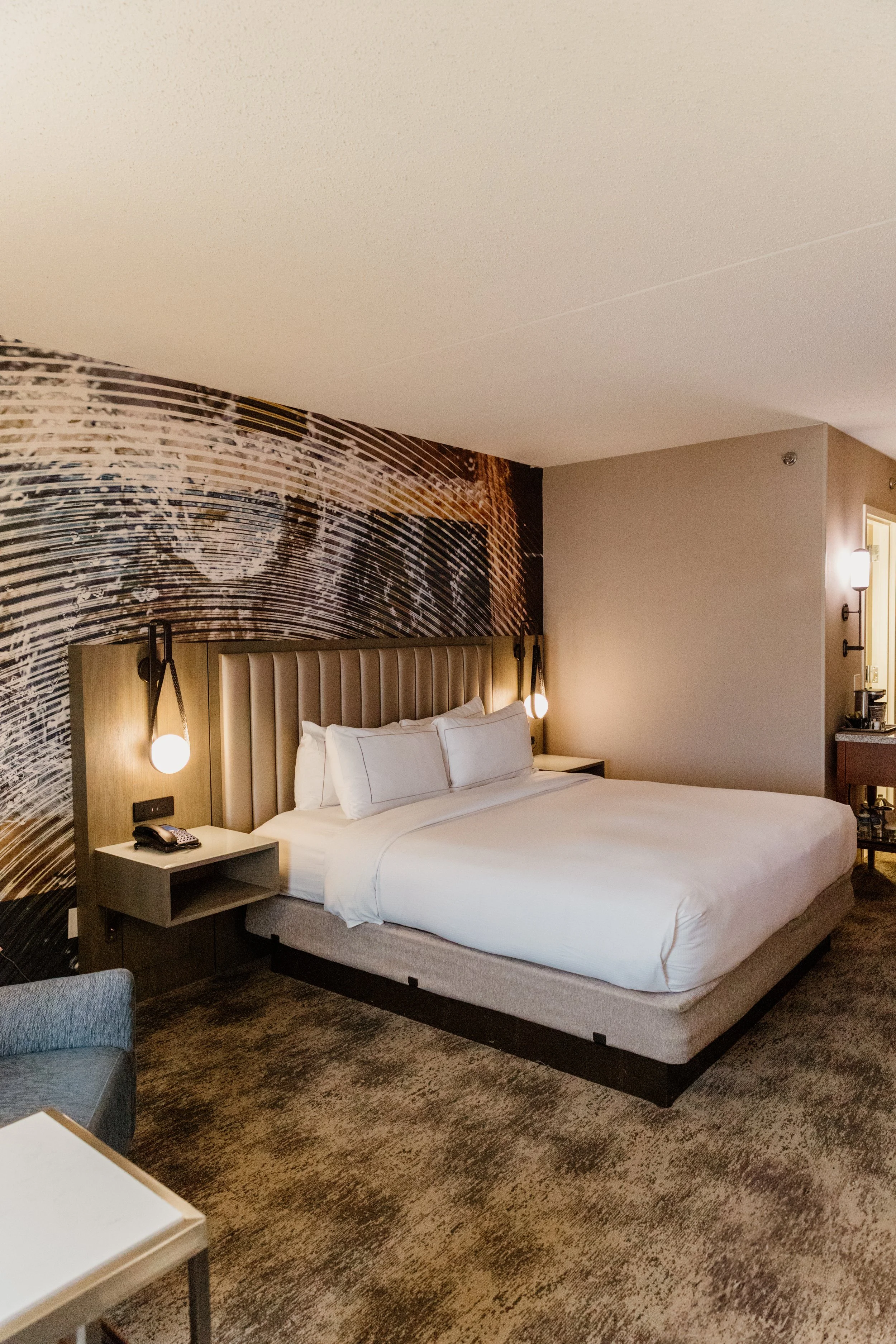 Luxury hotel room with king-sized bed, decorative headboard, wall-mounted lamps, patterned accent wall, and neutral-colored walls and carpet.