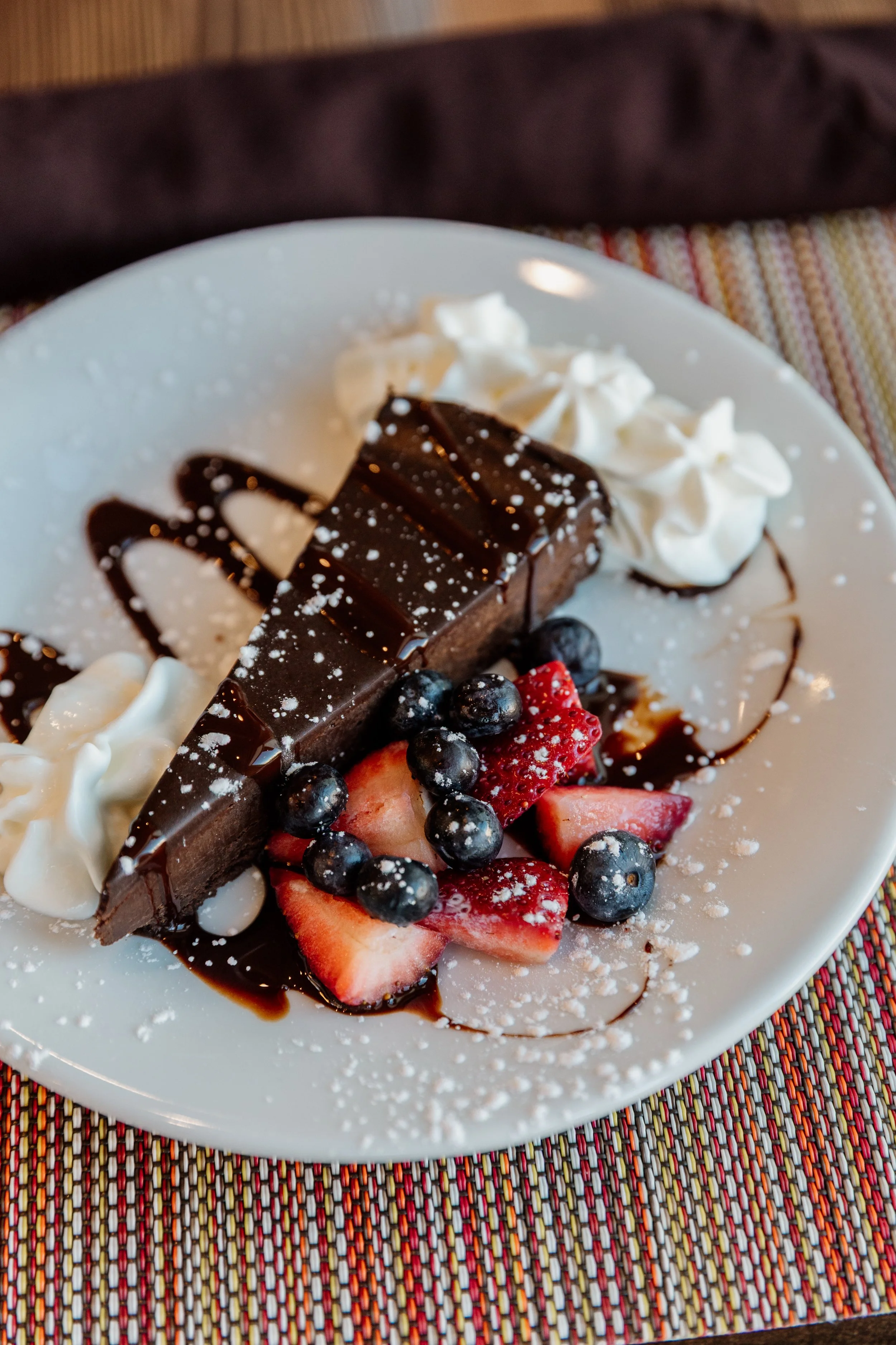 A dessert plate with a slice of chocolate cake, fresh strawberries, blueberries, whipped cream, a drizzle of chocolate sauce, and powdered sugar, on a colorful woven placemat.