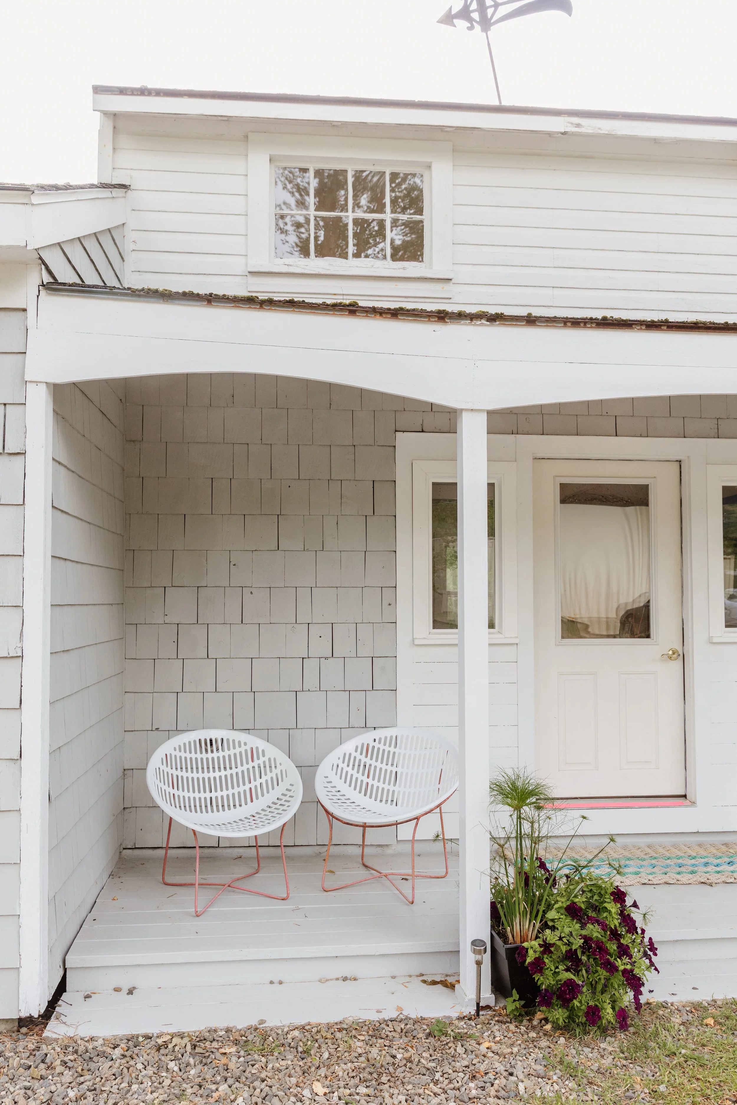 White porch with two modern chairs, potted plants, and a door with curtains.