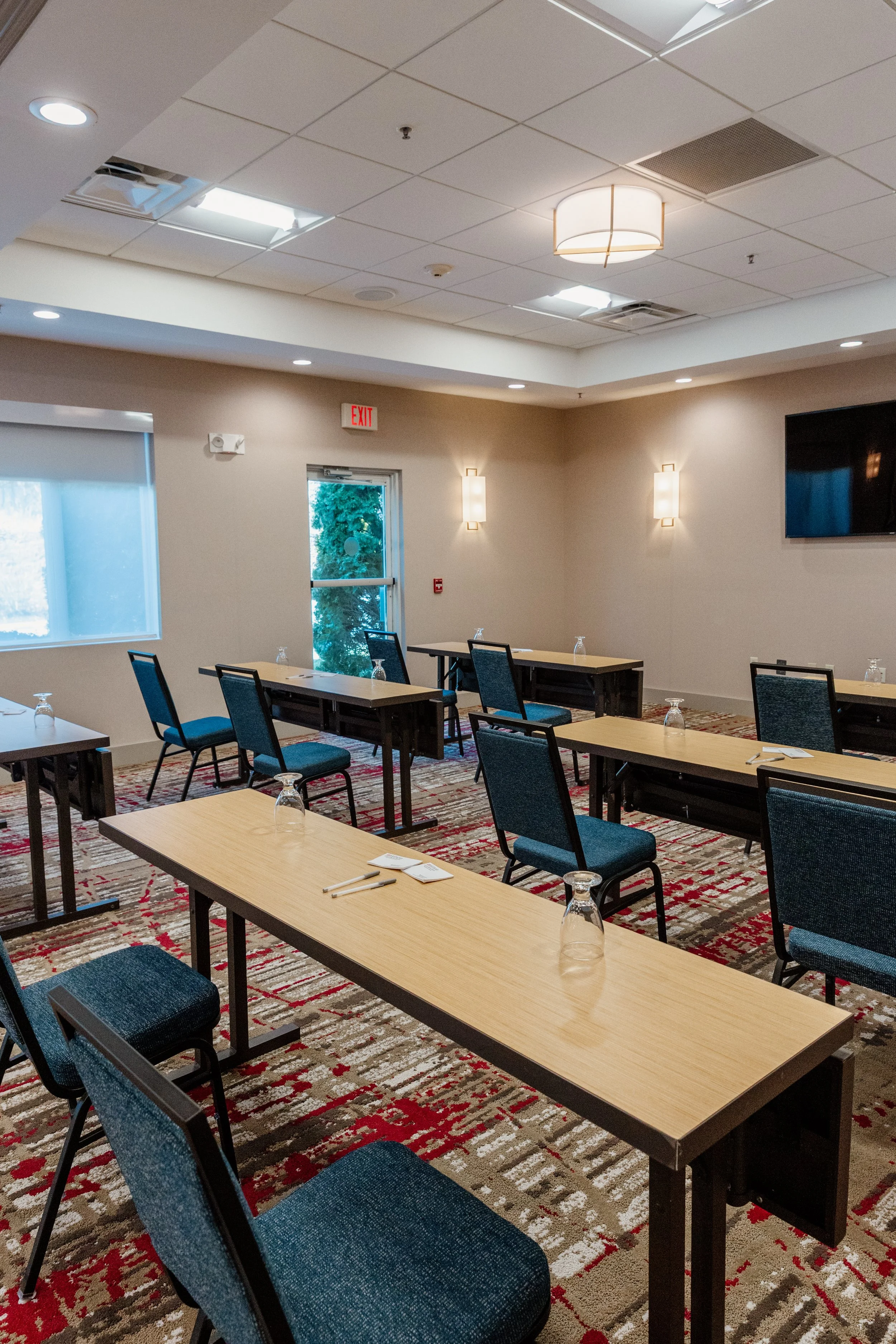 Empty conference room with tables, chairs, glasses, and writing utensils, illuminated by wall-mounted and ceiling lights, with a wall-mounted TV, window, and door.