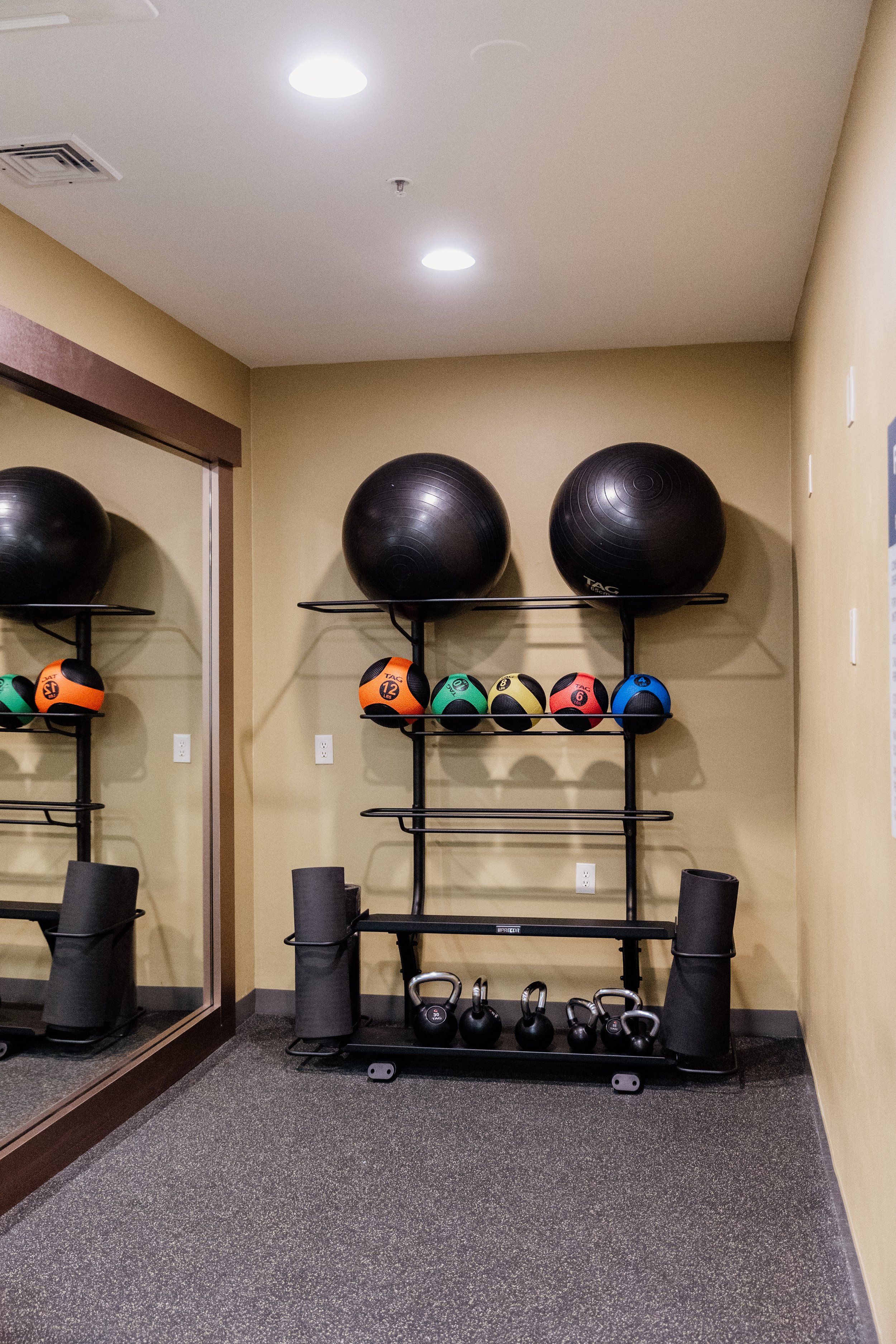A wall with exercise equipment, including large black exercise balls, colorful medicine balls, foam rollers, and kettlebells on a black metal rack, in a gym or fitness room.
