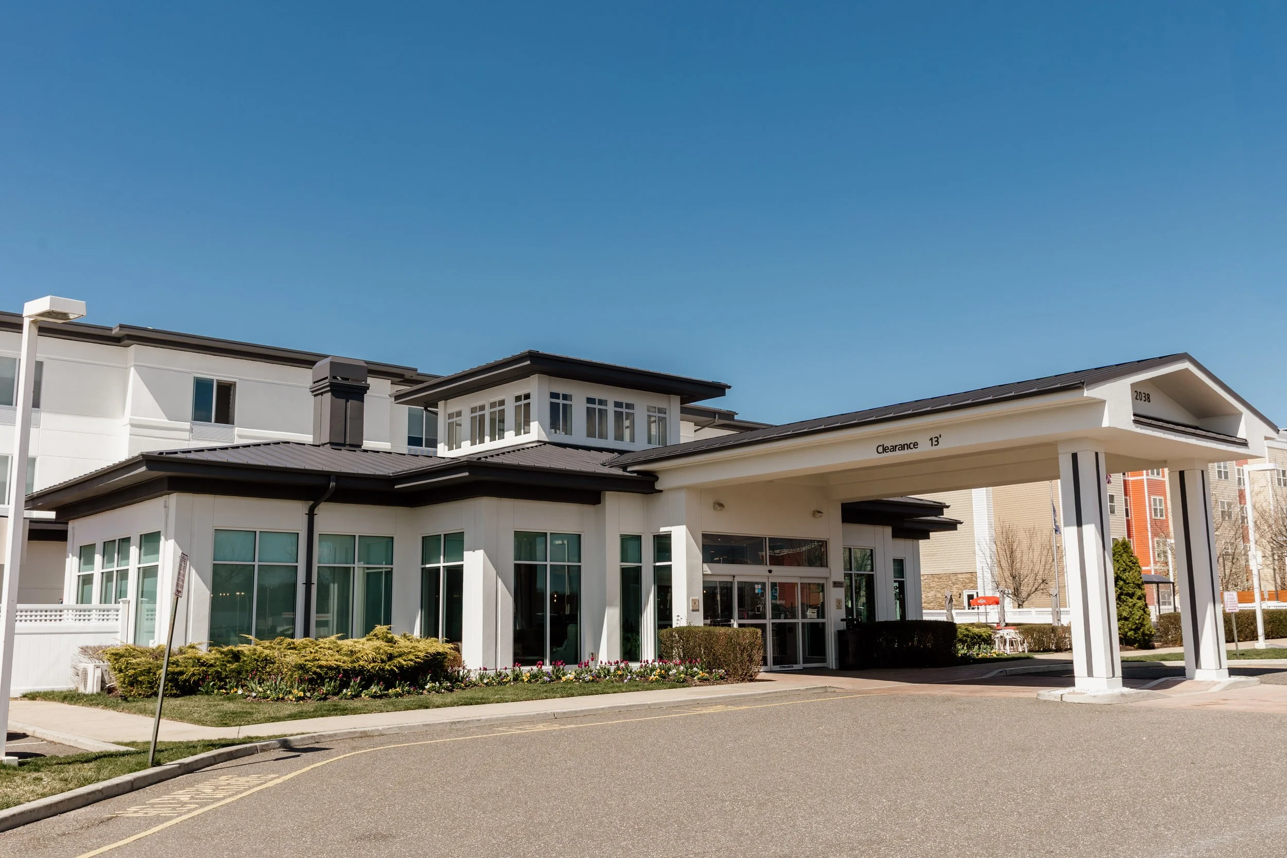 Exterior view of a modern residential building with a drive-thru covered entrance, clear blue sky, neatly landscaped garden with shrubs and flowers.