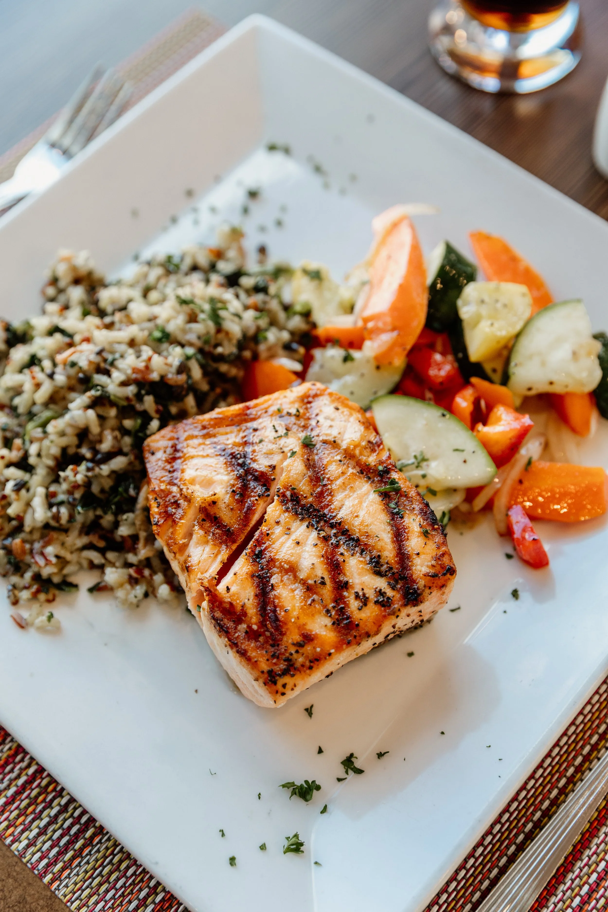 Grilled salmon fillet served with rice pilaf and steamed vegetable medley on a white square plate.