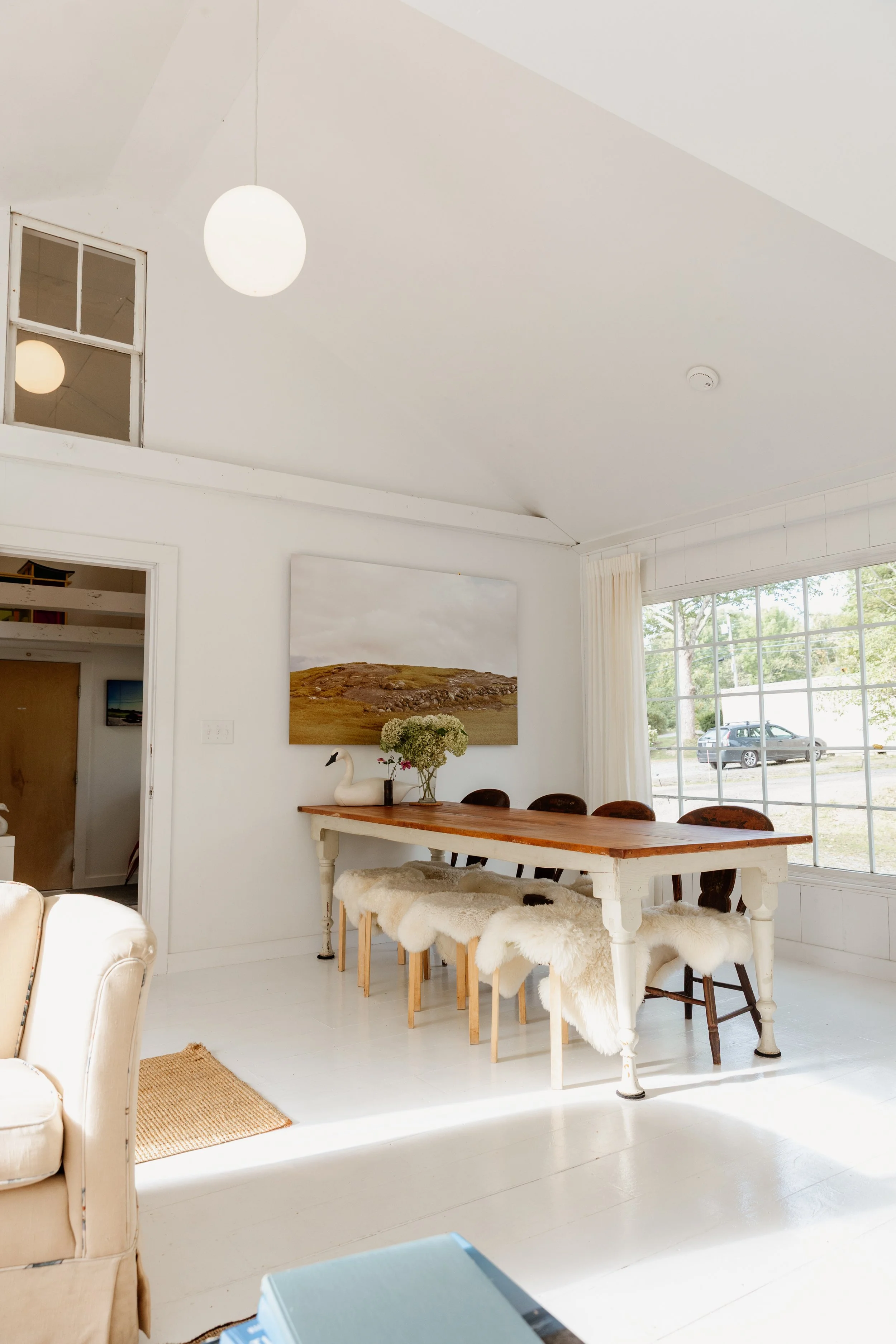 Bright dining room with a wooden table, surrounded by four chairs with plush sheepskin covers, positioned in front of large window with curtains, and decorated with a vase of flowers and a swan-shaped sculpture. A large landscape photograph hangs on 