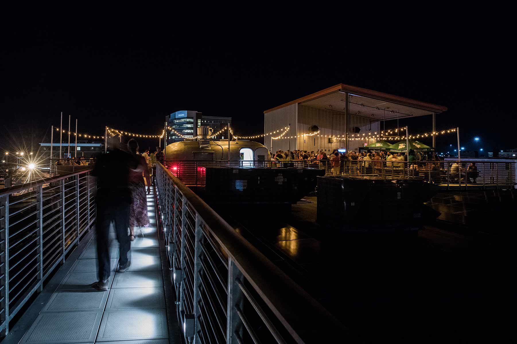 People walking a catwalk to a rooftop bar with string lights and a crowd of people