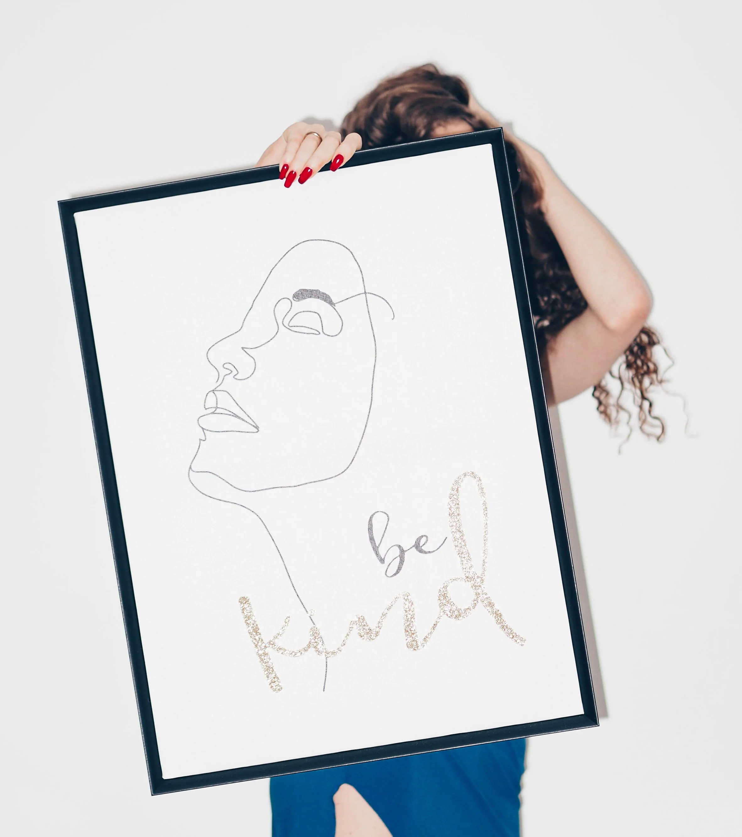 A person with curly hair holding a framed minimalist line drawing of a woman's face and the words "be kind" on a white background.
