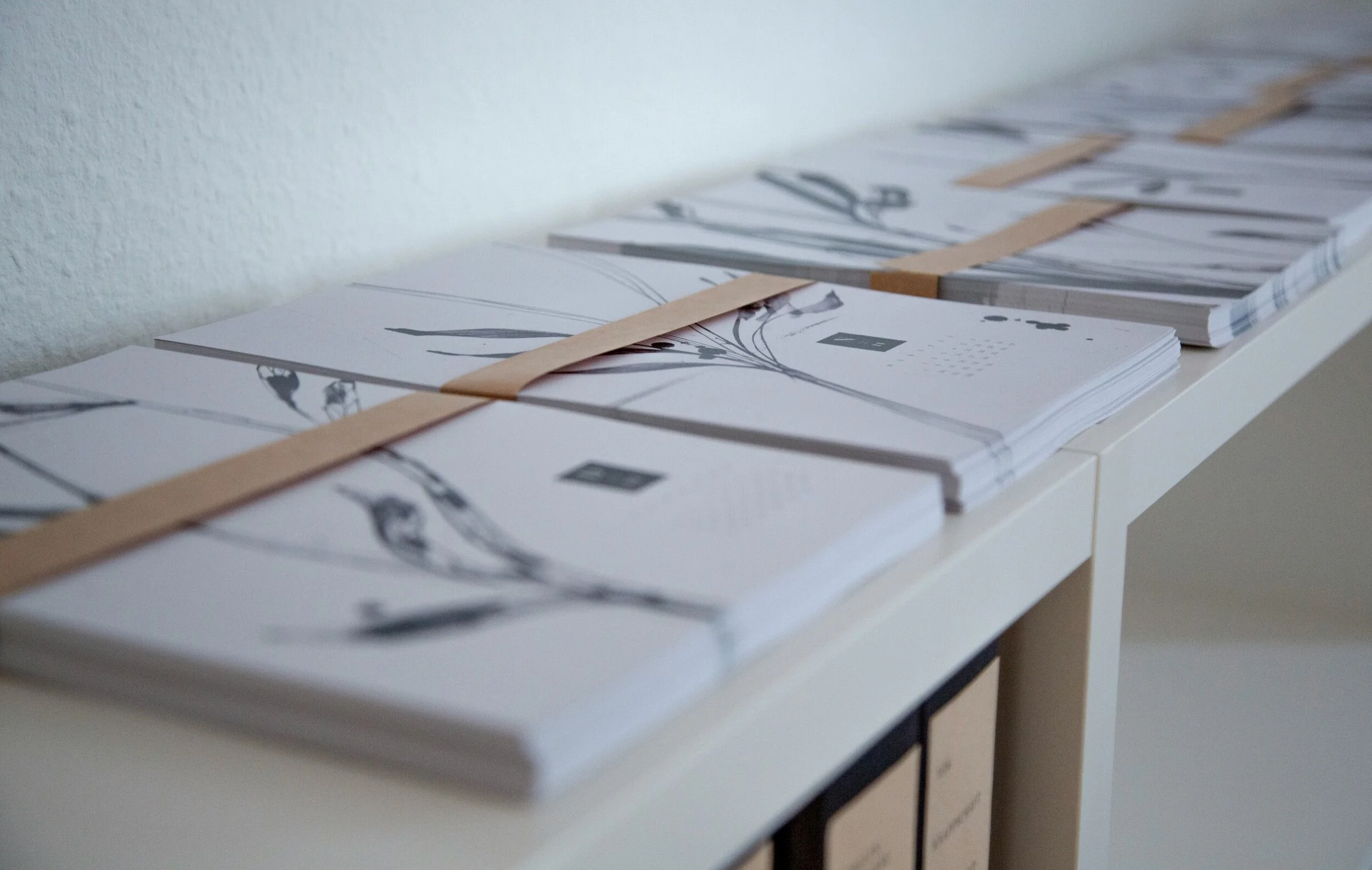 Stacks of white paper with black floral illustrations, held together with brown paper straps, on a white shelf.