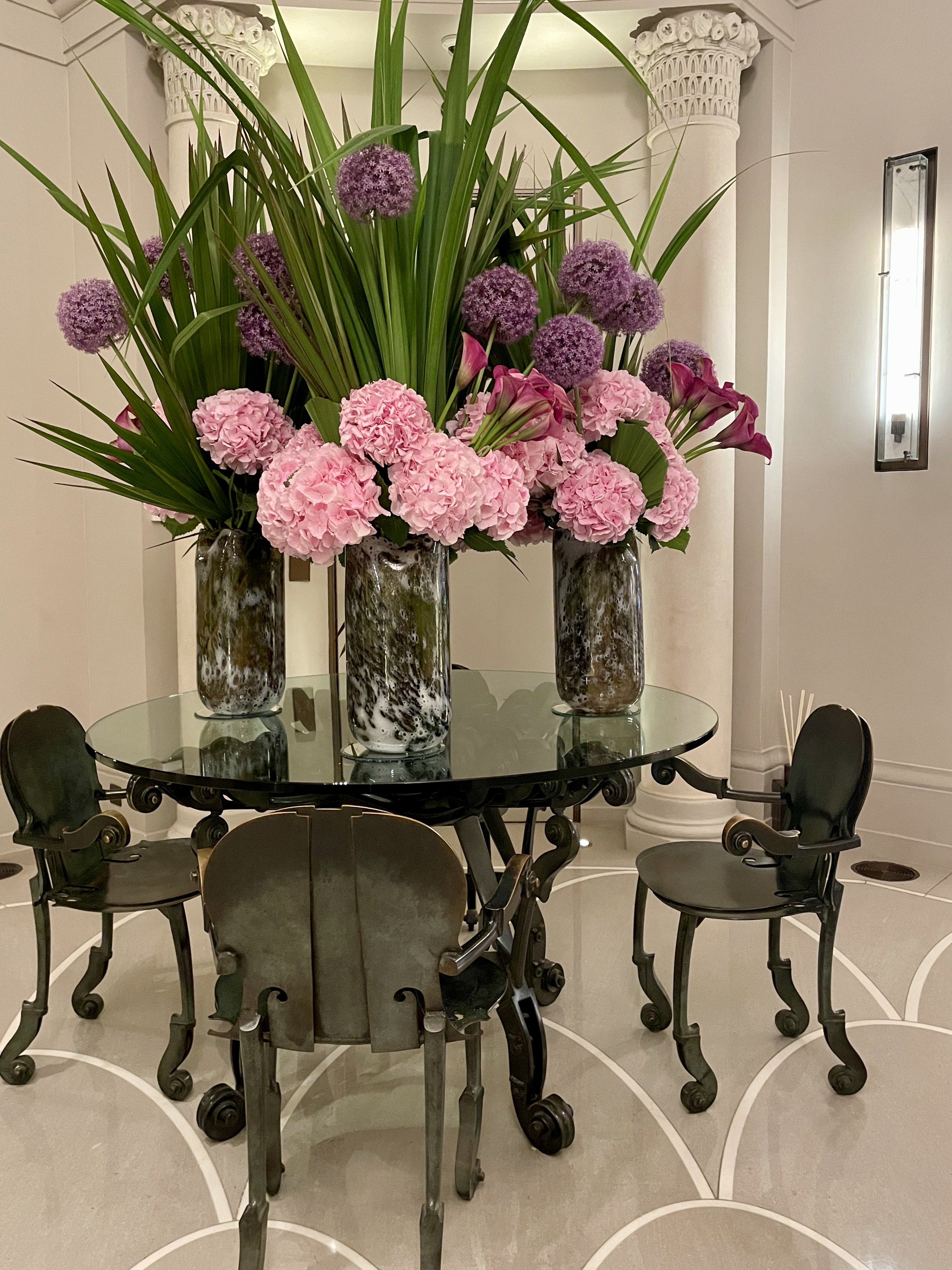 A luxury Palm Beach European philosophy dining space with a vintage deco metal table and chairs is graced by vibrant hydrangeas and calla lilies in three vases. Classic architecture has beautiful columns.