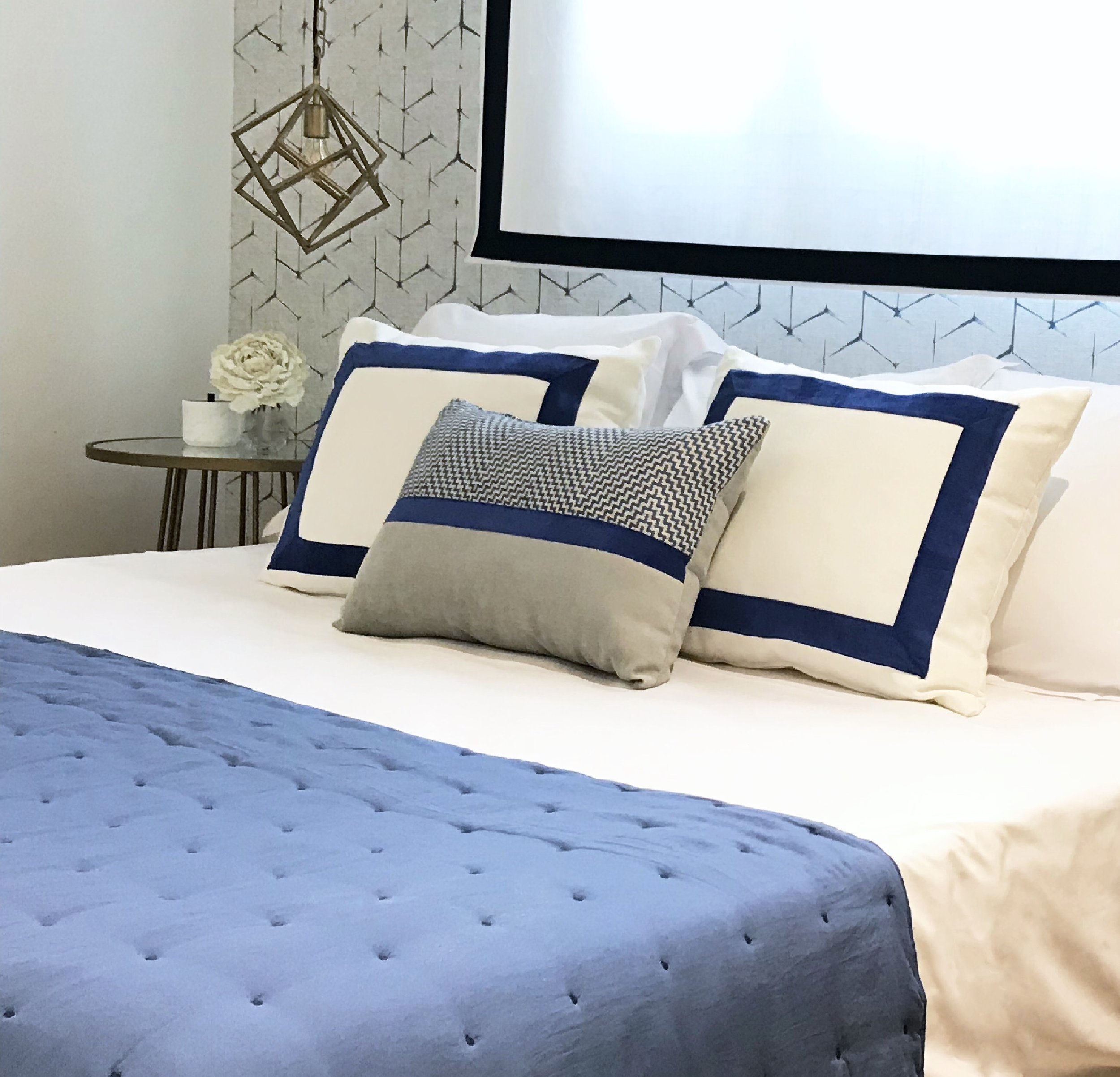 Image of professionally styled bed in blues and off white tones with graphic wallpaper and hanging gold, dynamic light.