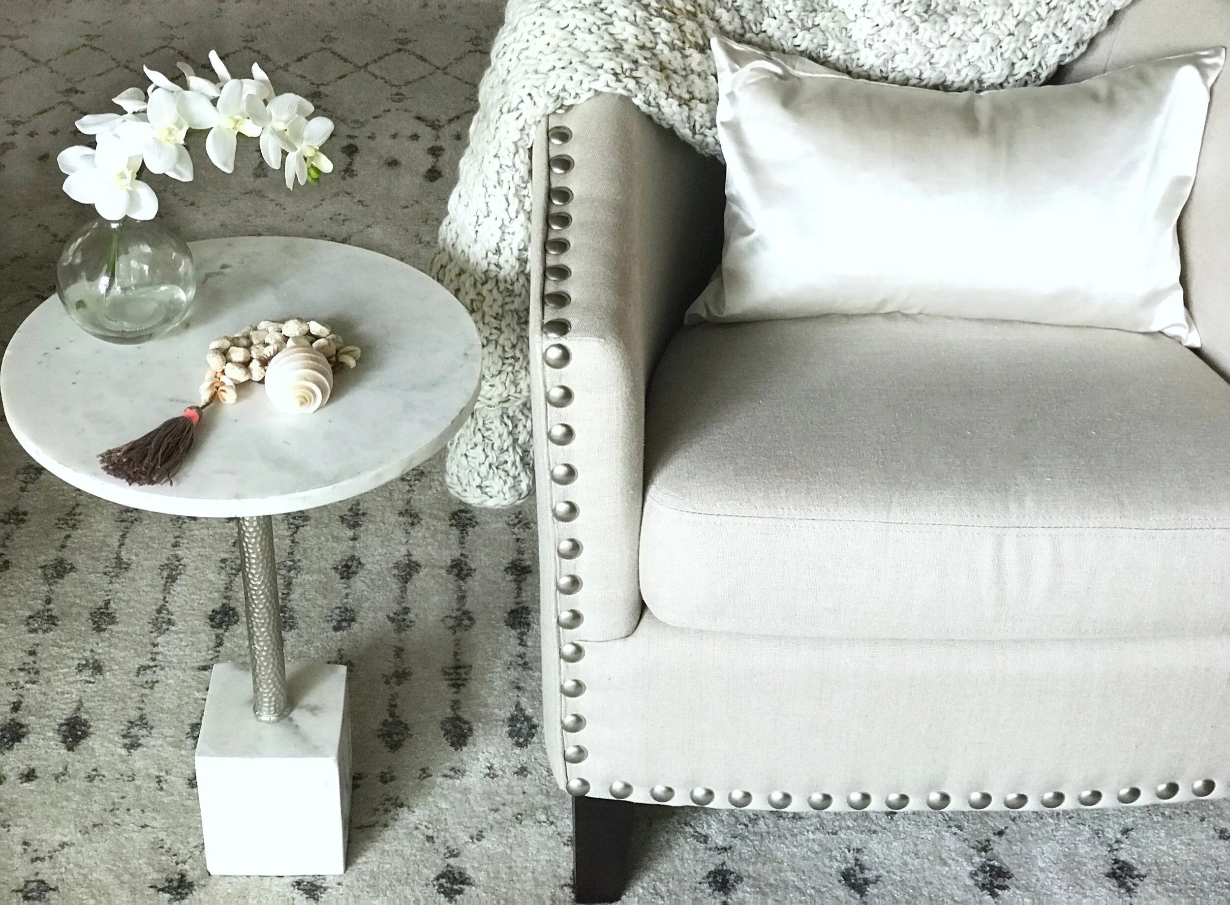 Luxury living by Domaine Luxe shows a cozy spot  for reading with a linen club chair detailed with silver nail heads, with a plush knit throw, next to a round marble and steel side table, graced with an orchid and shells, on a patterned ivory rug.