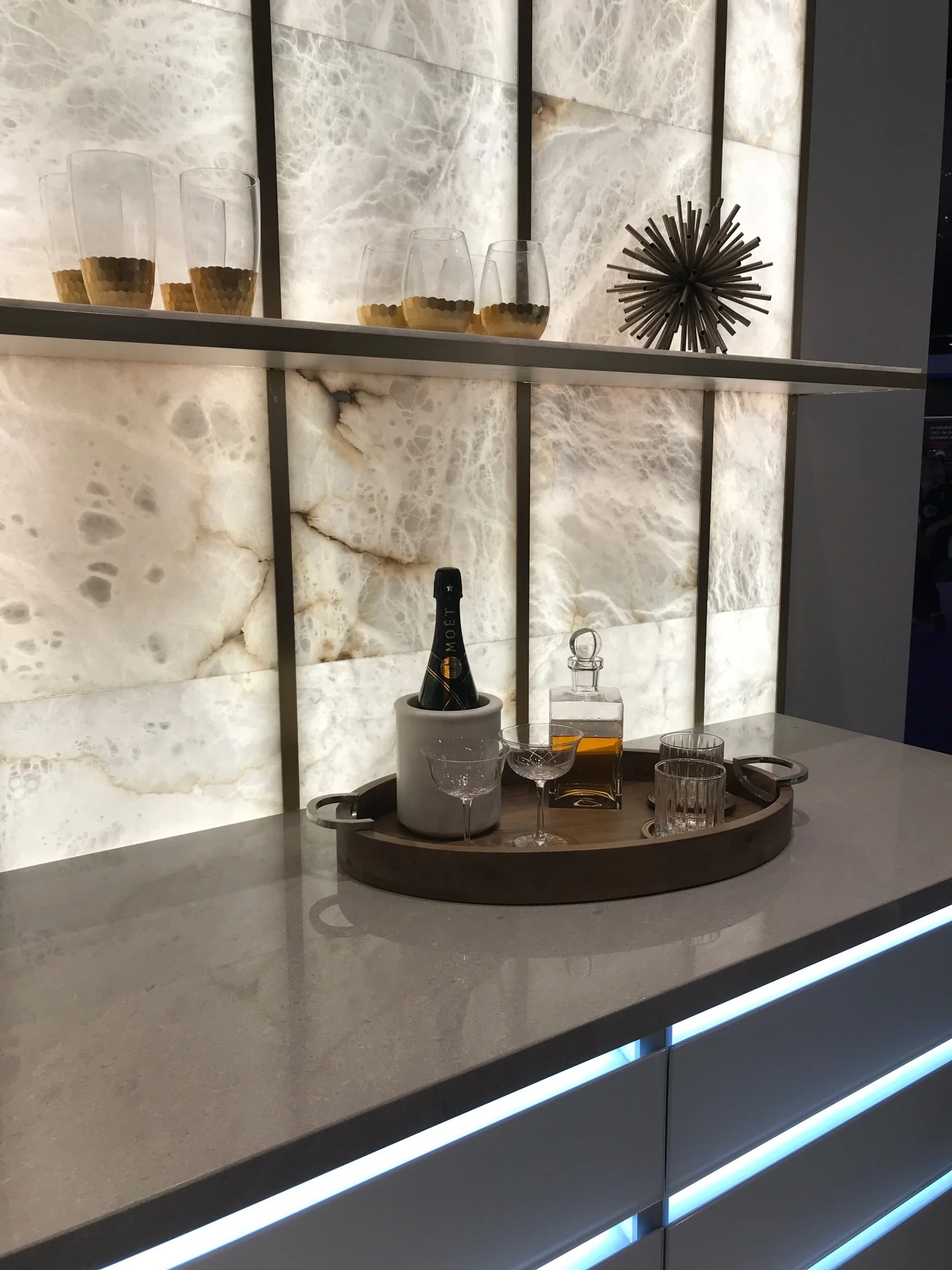 Architectural finishes for interior design in Palm Beach shows a bar with stone backsplash with lighting from behind, set with bar glasses and accessories.