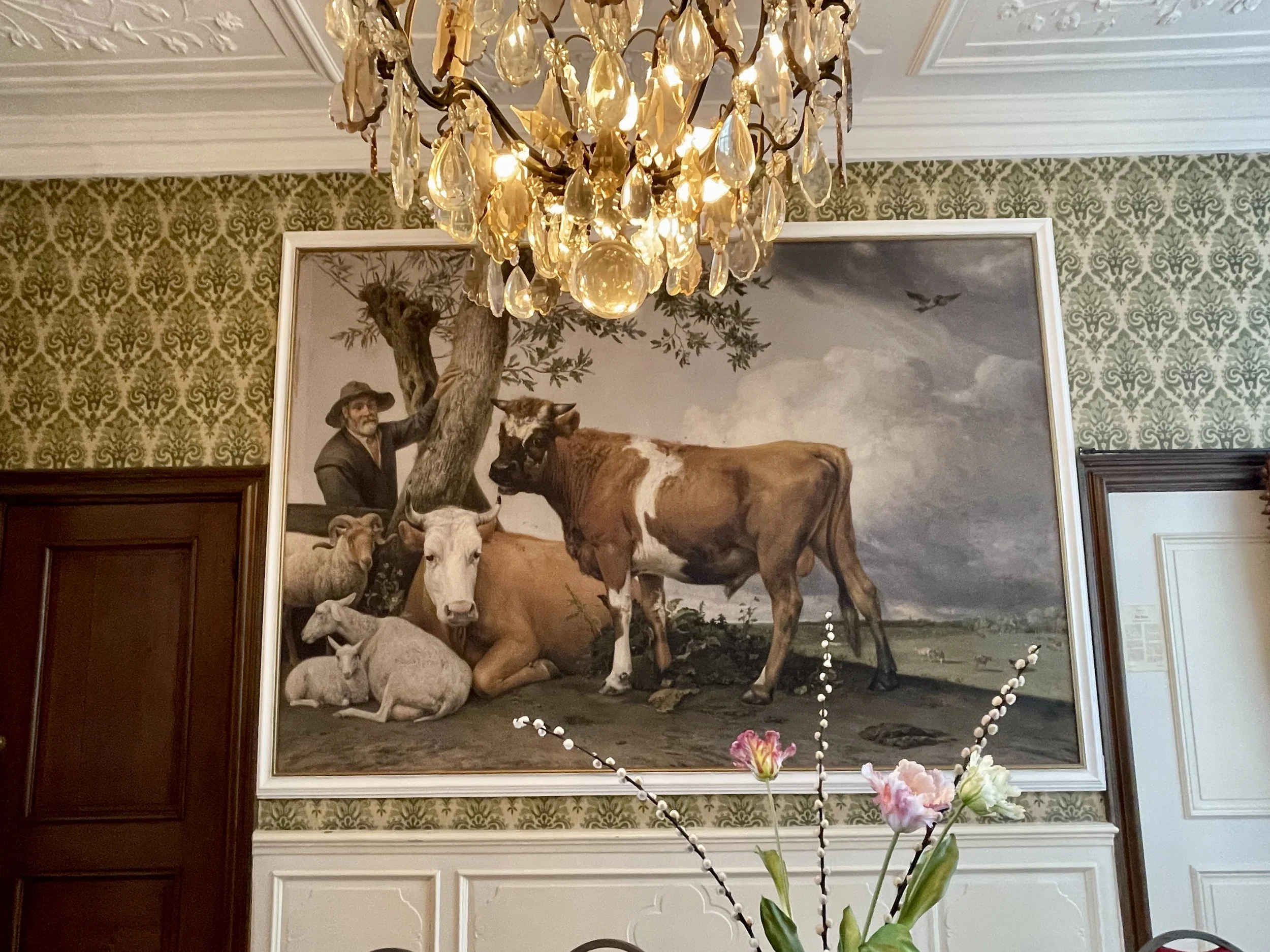 Historic Dutch dining room with large pastoral painting, crystal chandelier, and patterned walls illustrating quiet European luxury