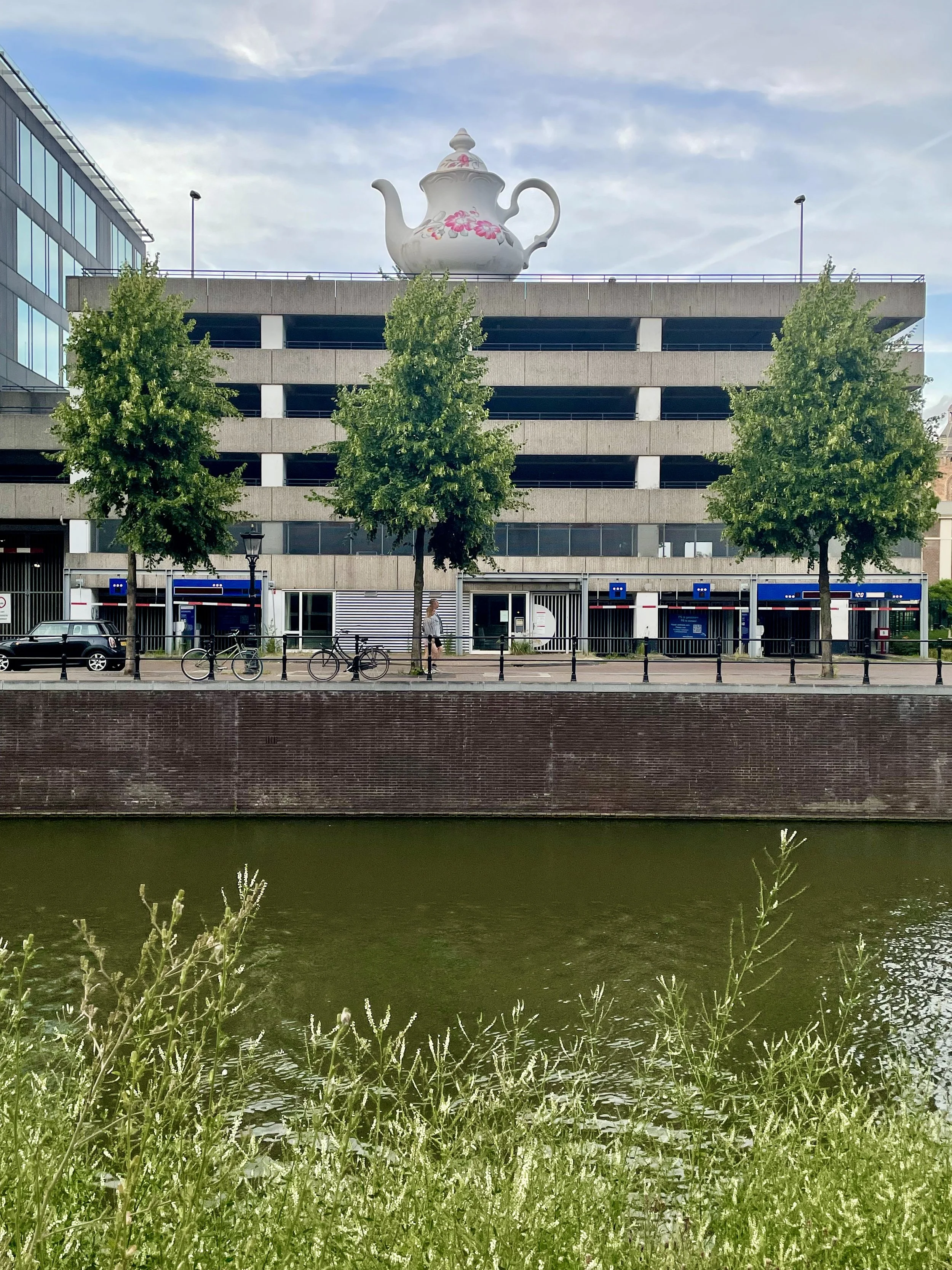 A whimsical teapot in an antique style donned with flowers perches on top of a parking garage along side the canal in Utrecht.