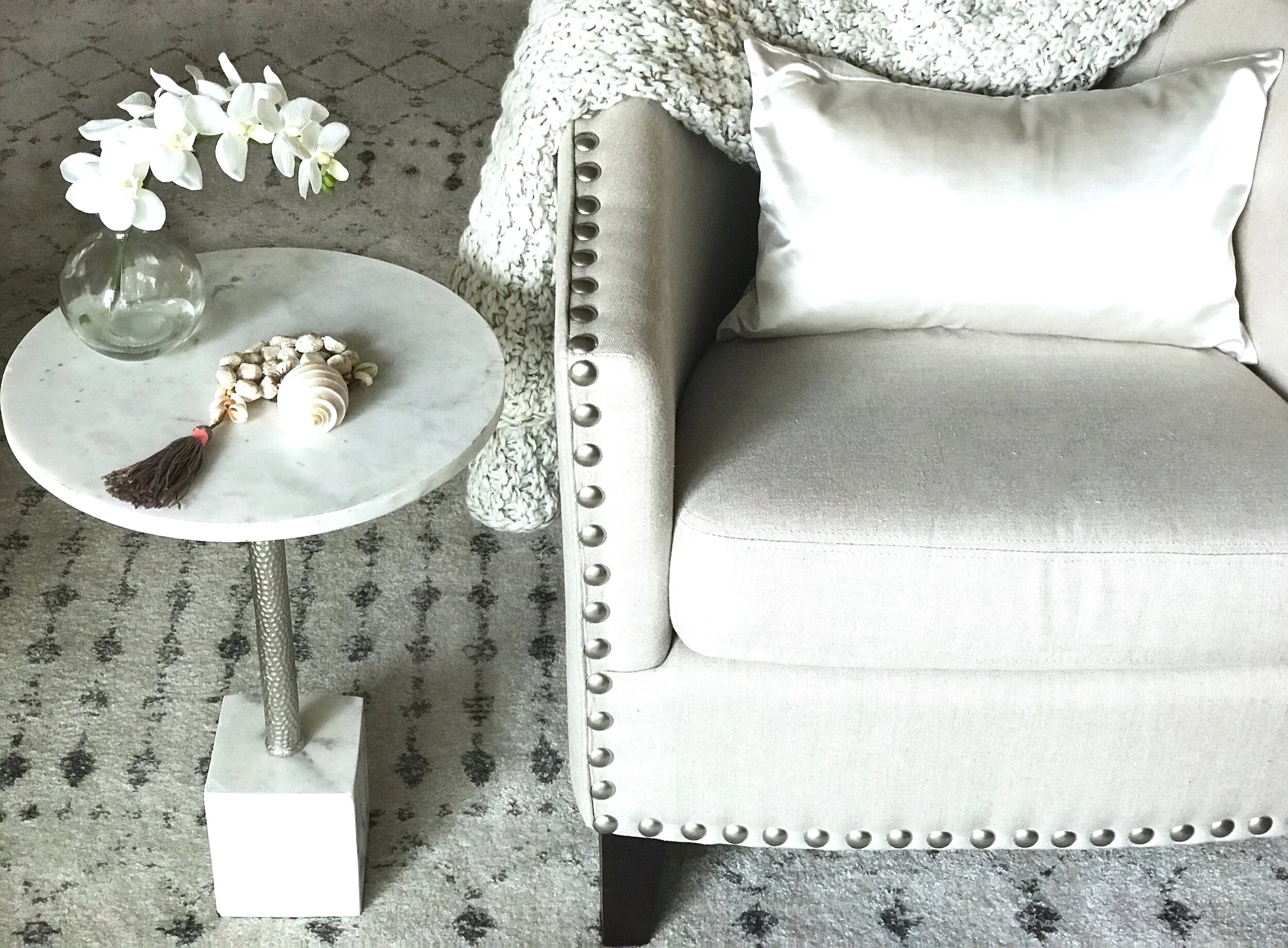 Luxury living by Domaine Luxe shows a cozy spot  for reading with a linen upholstered club chair detailed with silver nail heads, a plush knit throw, a round marble steel side table, graced with an orchid and shells, on a patterned ivory rug.