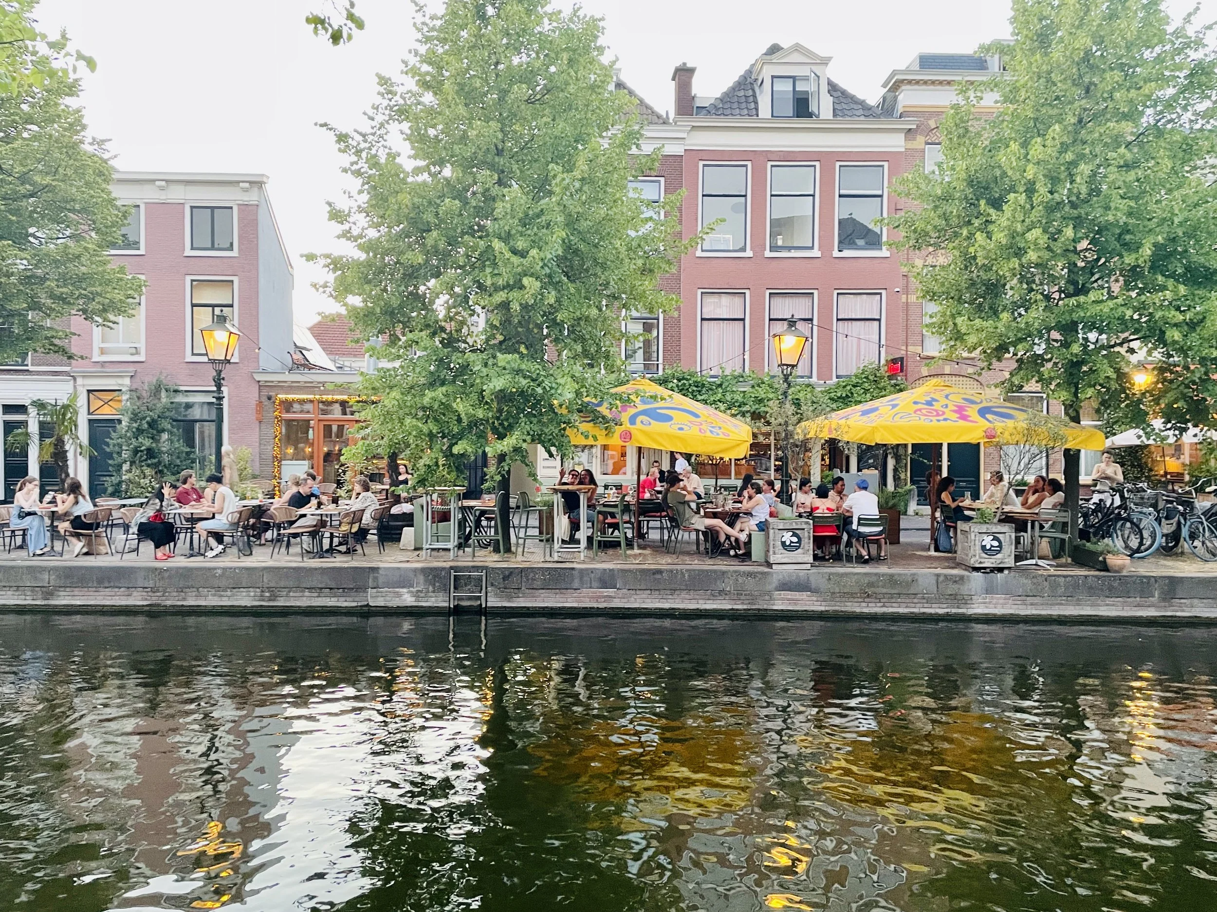 Canal-side café in The Hague with people dining outdoors under trees and yellow umbrellas beside the water.