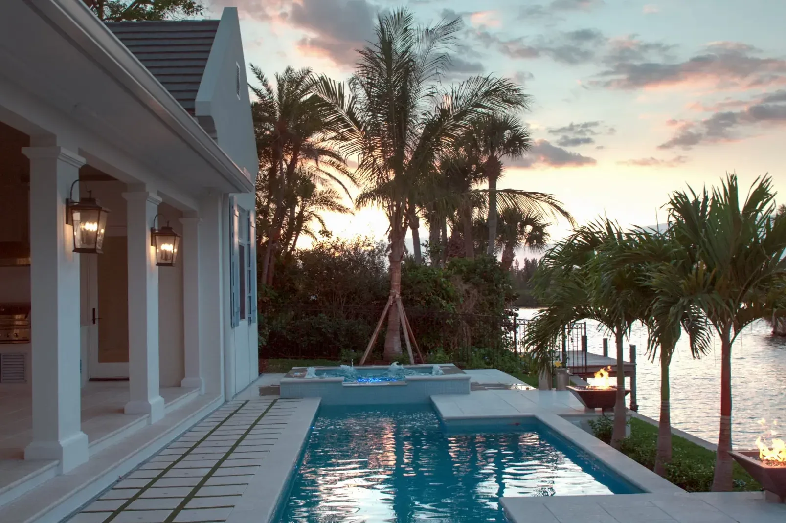 Waterfront Palm Beach pool and spa at sunset, fire pit going, with lights illuminating the covered loggia columns facing the pool.