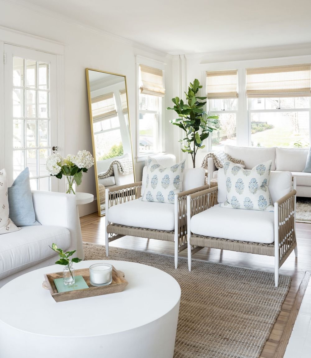 Light-filled coastal living room with white sofa, woven armchairs, jute rug, and blue patterned pillows, and lush plants and hydrangea flowers in vase.