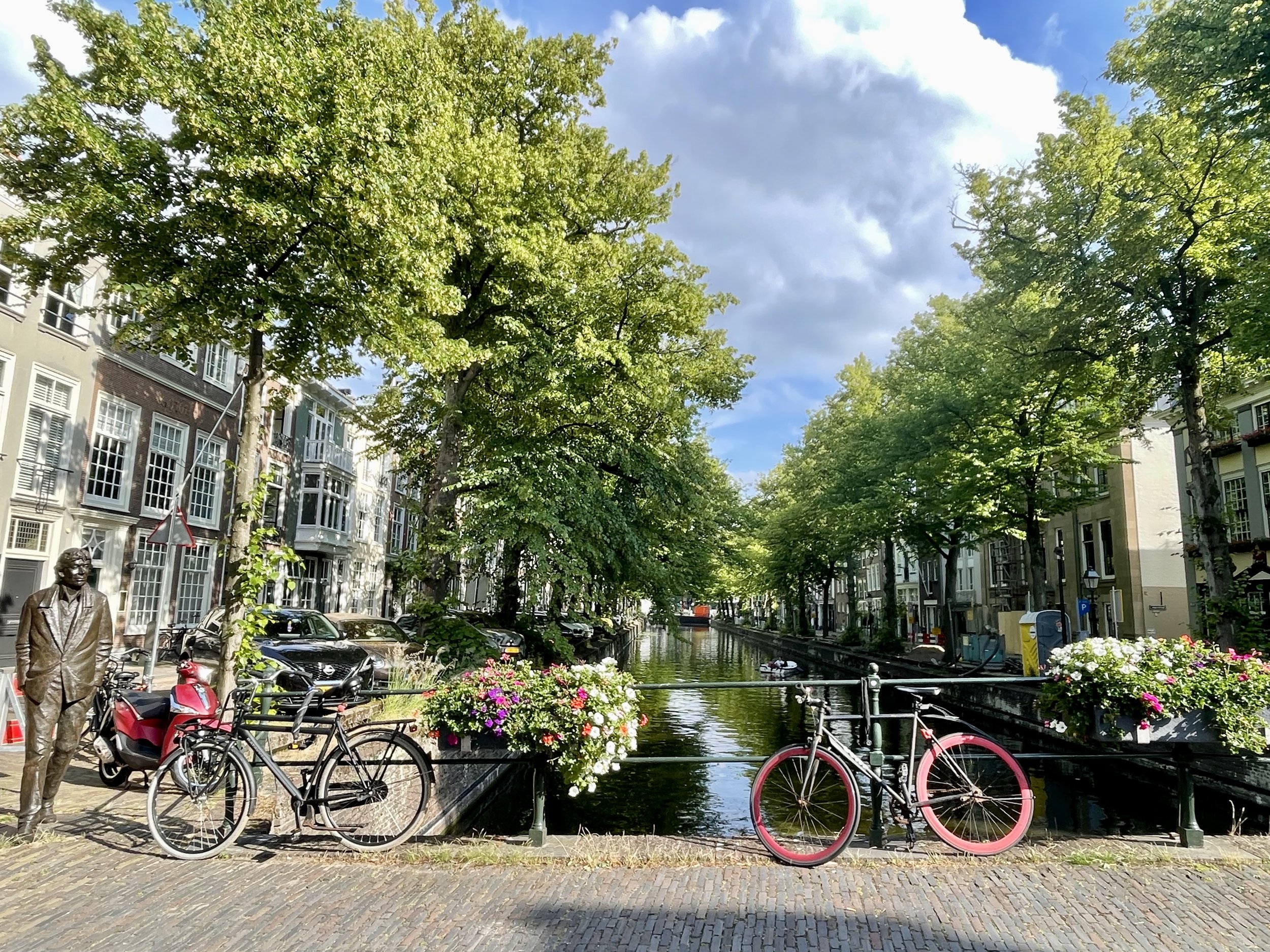 Tree-lined Dutch canal with bicycles on a bridge, flower boxes, and historic townhouses under a bright sky.