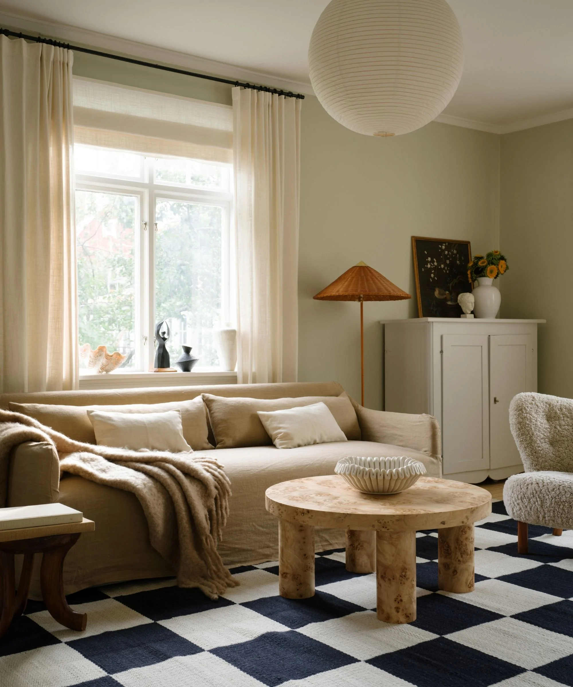 European-inspired sitting room with linen drapery, neutral sofa, burlwood round coffee table, and filtered natural light across a graphic black and white soft checkered rug.