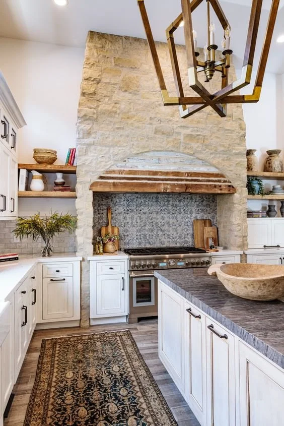 Daylight grounds this European-inspired kitchen with white cabinetry, stone and wood range hood, and brass chandelier.